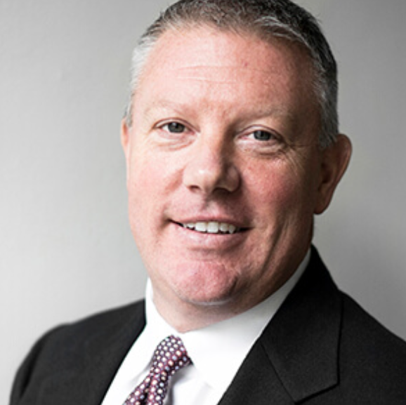 Joel Rohrs, A professional man with light skin, short gray hair, blue eyes, wearing a black suit, white shirt, and patterned tie, smiling at the camera against a plain background.