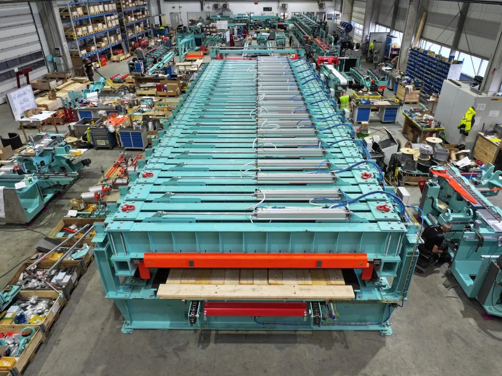 Large industrial manufacturing facility with a long, row of teal-colored CLT machinery central to the factory. Workers and various equipment surround the machinery, and shelves with parts line the walls.