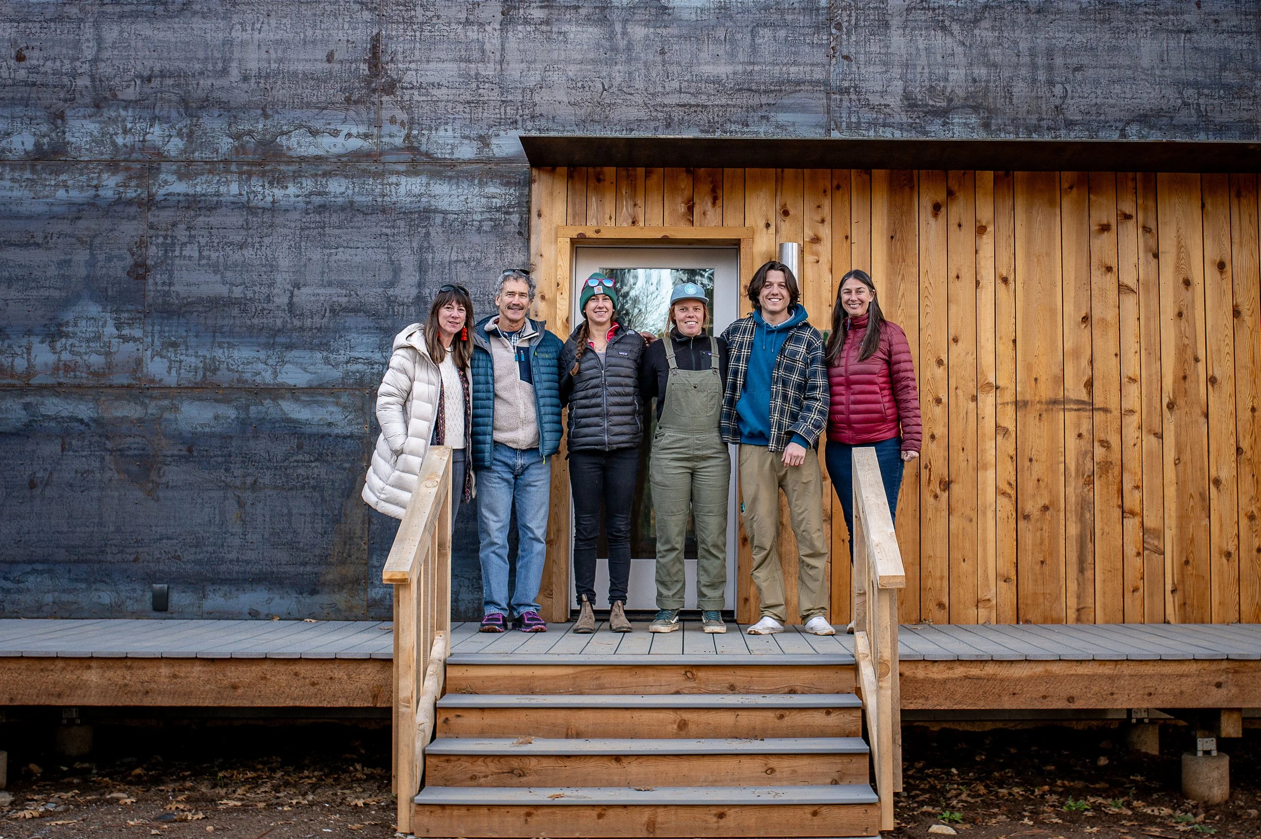 Group of six people standing on CLT wooden stairs in front of a CLT home with a mix of wood and steel siding, posing for photo outdoors.