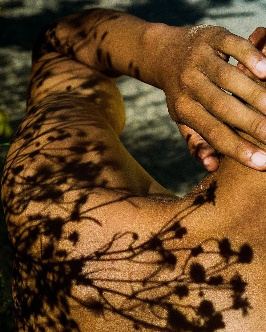 Close-up of a person with a shadow of leaves and flowers cast on their skin, with one hand resting on their shoulder.