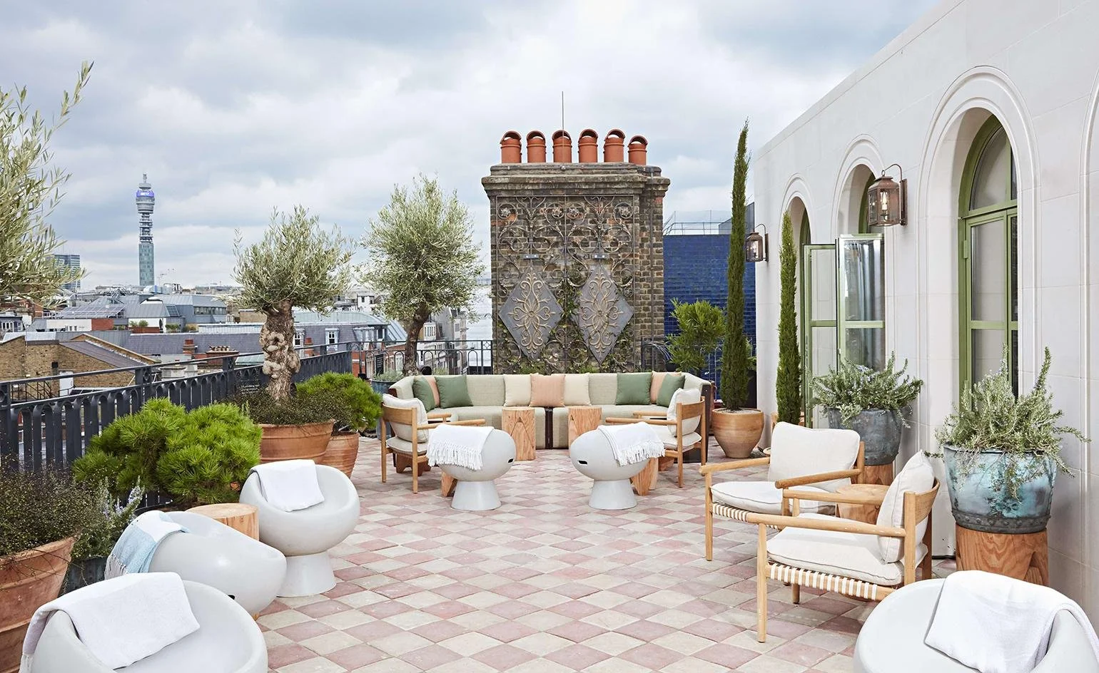 Rooftop terrace with modern outdoor furniture, potted plants, olive trees, and cityscape view, including a tall TV tower in the background.