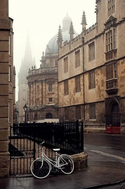 Empty street with historic stone buildings, a white bicycle parked against a fence, and a foggy, overcast atmosphere.