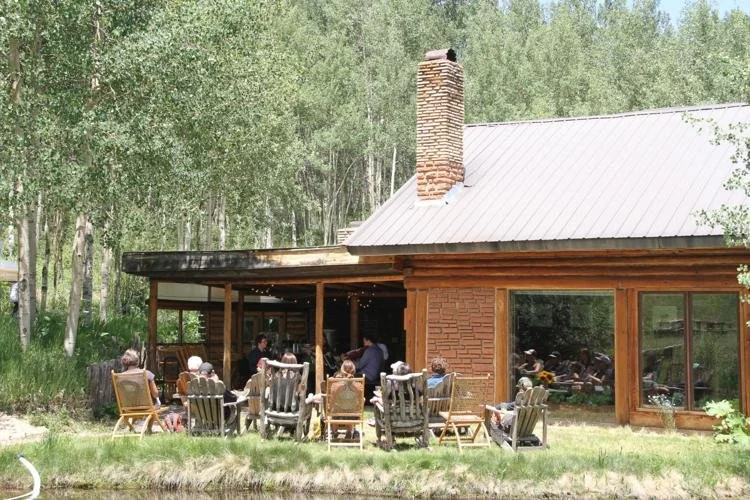 People enjoying a gathering on an outdoor patio of a rustic house surrounded by trees, with a brick chimney and large windows.