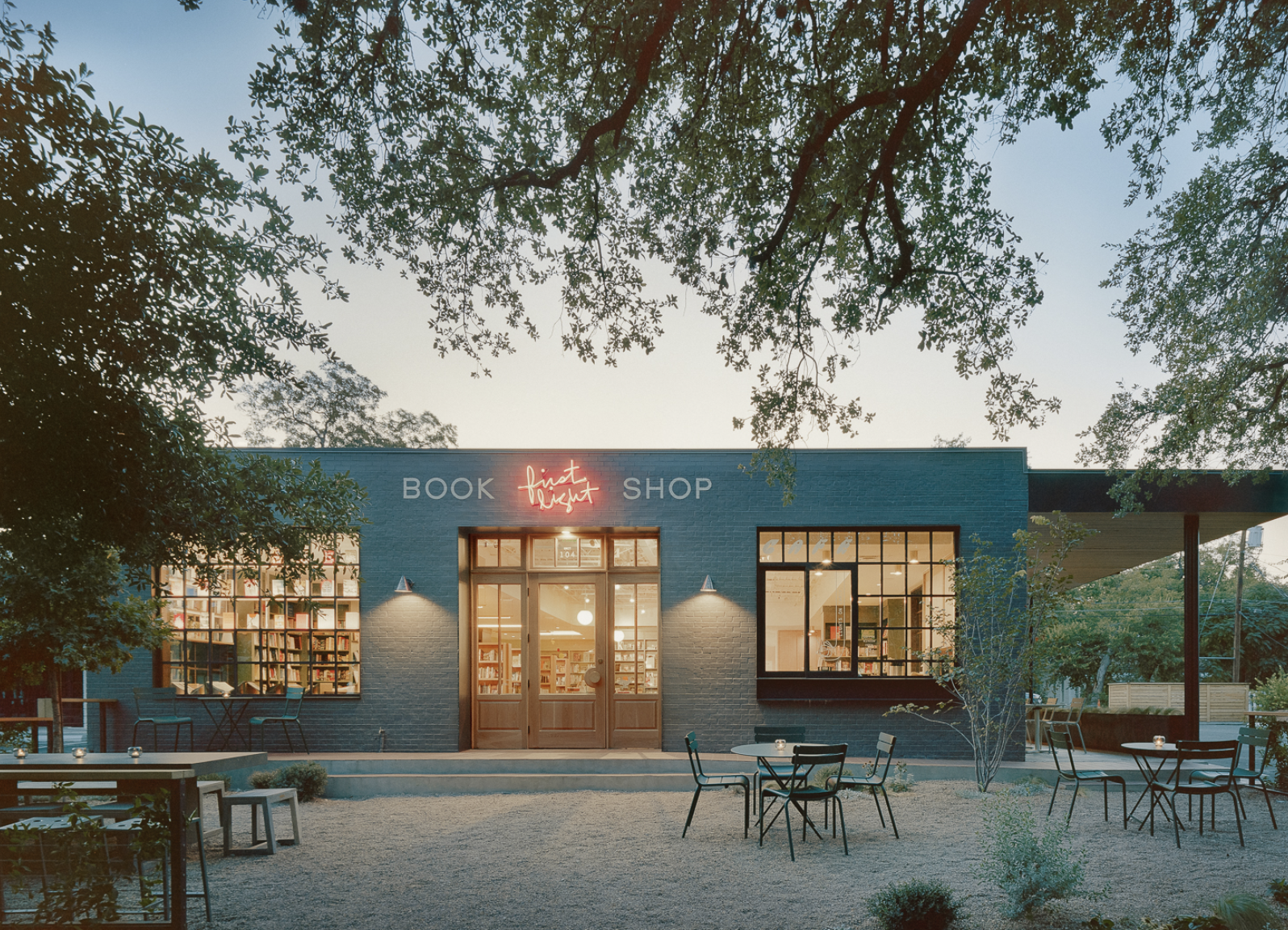 Exterior view of a bookstore called first light shop at dusk, with outdoor tables and chairs, large windows showing books inside, and trees framing the scene.