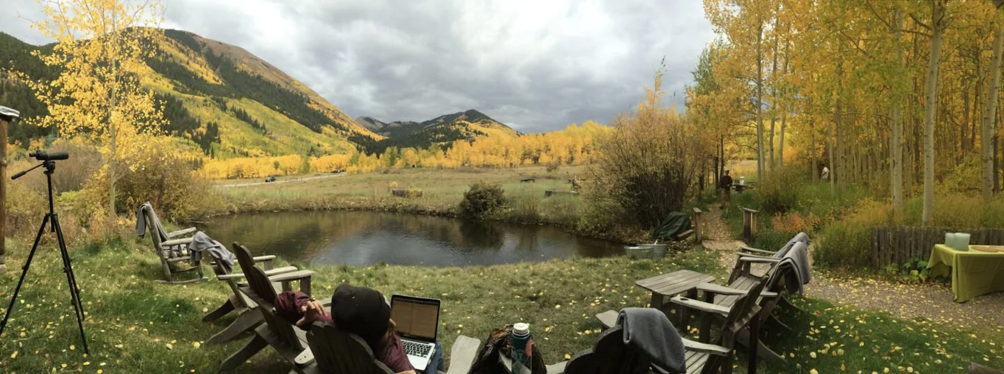 Outdoor scene with Adirondack chairs and a picnic table near a pond, surrounded by fall foliage and mountains under cloudy sky.