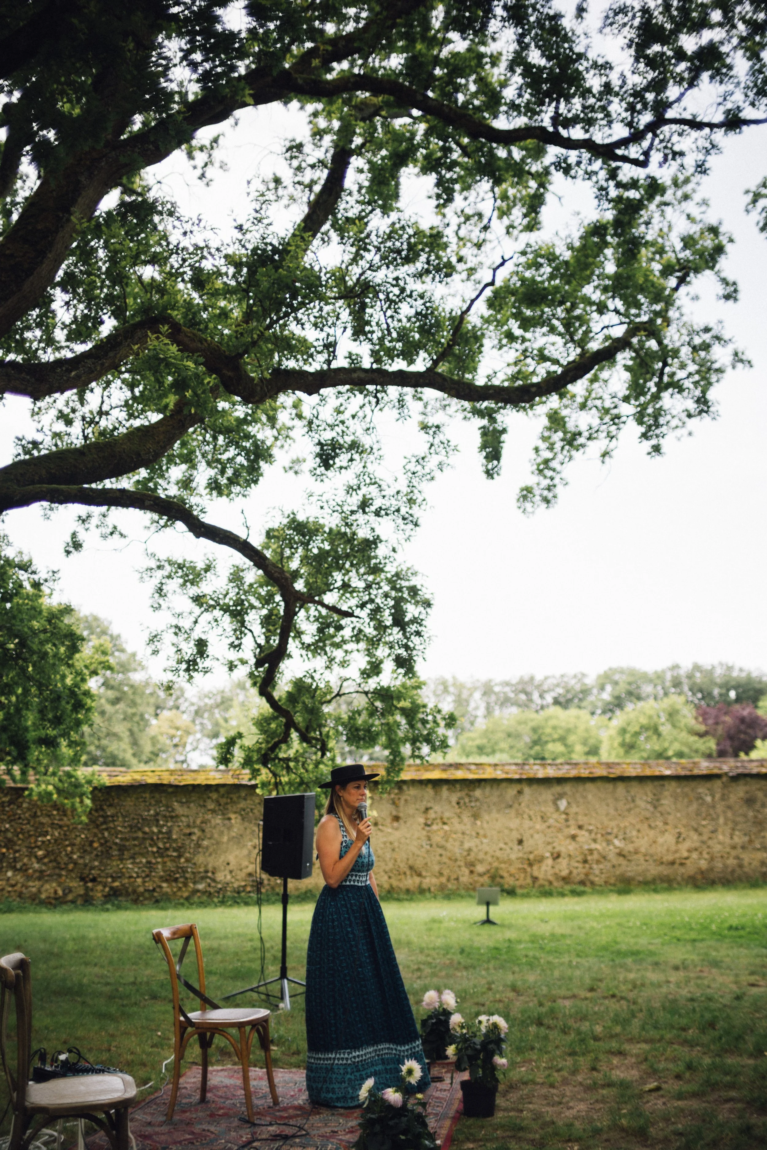A woman in a long dark dress and wide-brimmed black hat speaks into a microphone outdoors, standing on a small rug with potted flowers nearby, under a large tree with green leaves, behind an old stone wall.