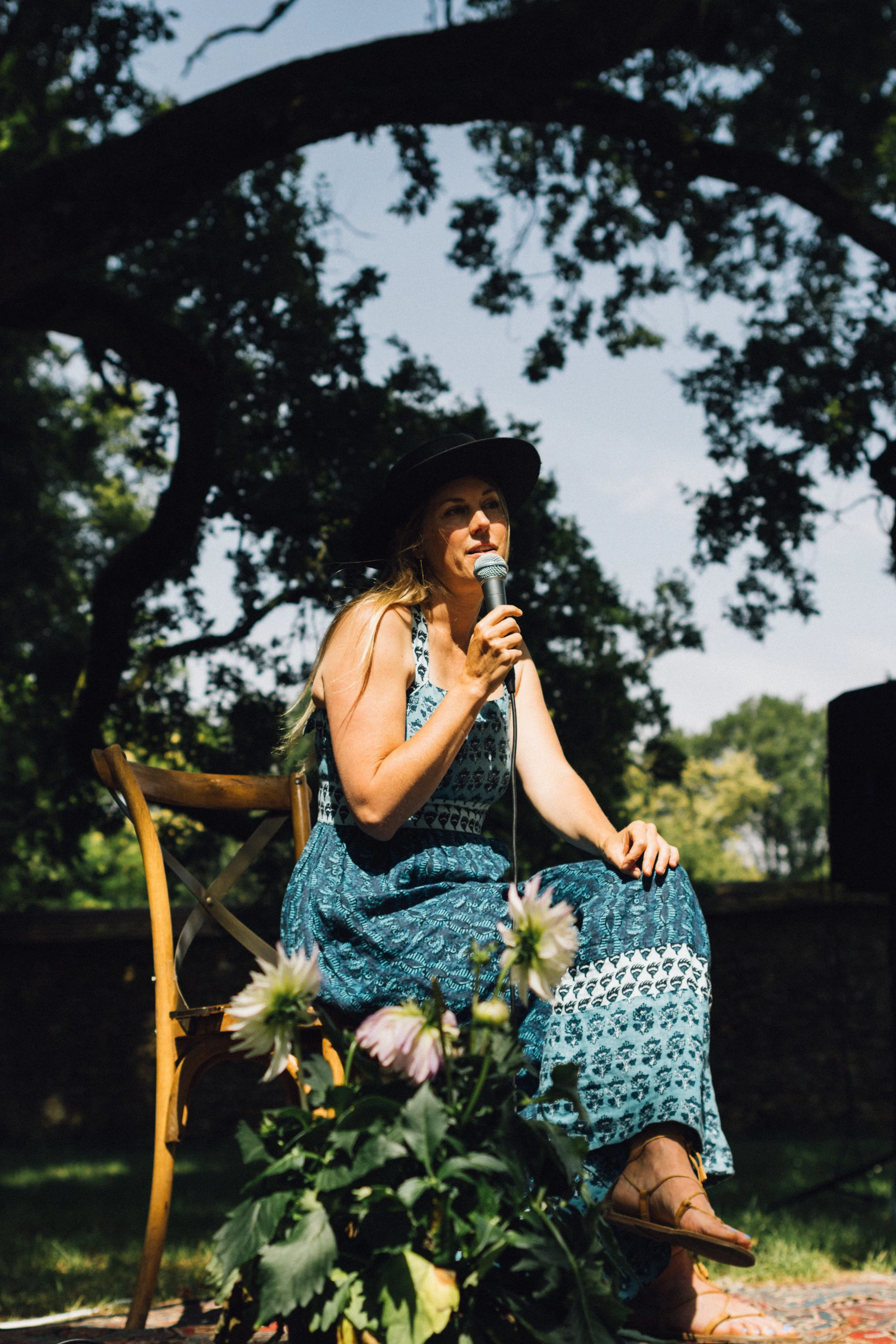A woman sitting on a wooden chair outdoors, holding a microphone, wearing a wide-brimmed hat and a blue patterned dress, with flowers in front of her and a giant old oak tree in the background.