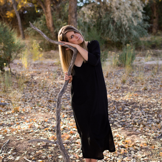 A woman in a black dress leaning her head on a large tree branch in a wooded area during daytime.