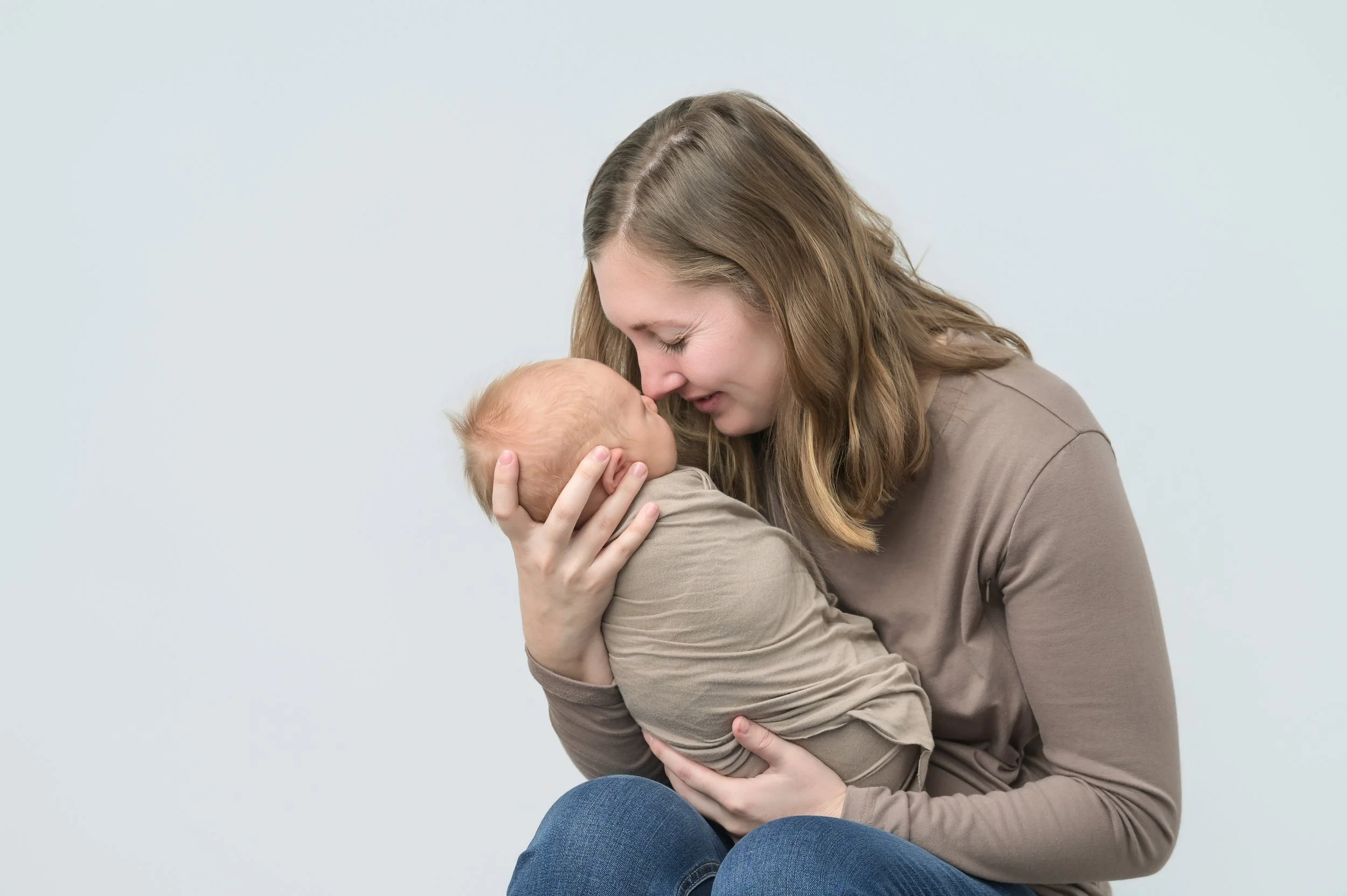A woman holding a baby close to her face, touching foreheads, with a gentle smile, against a plain light background. This is a newborn portrait session with Mother.