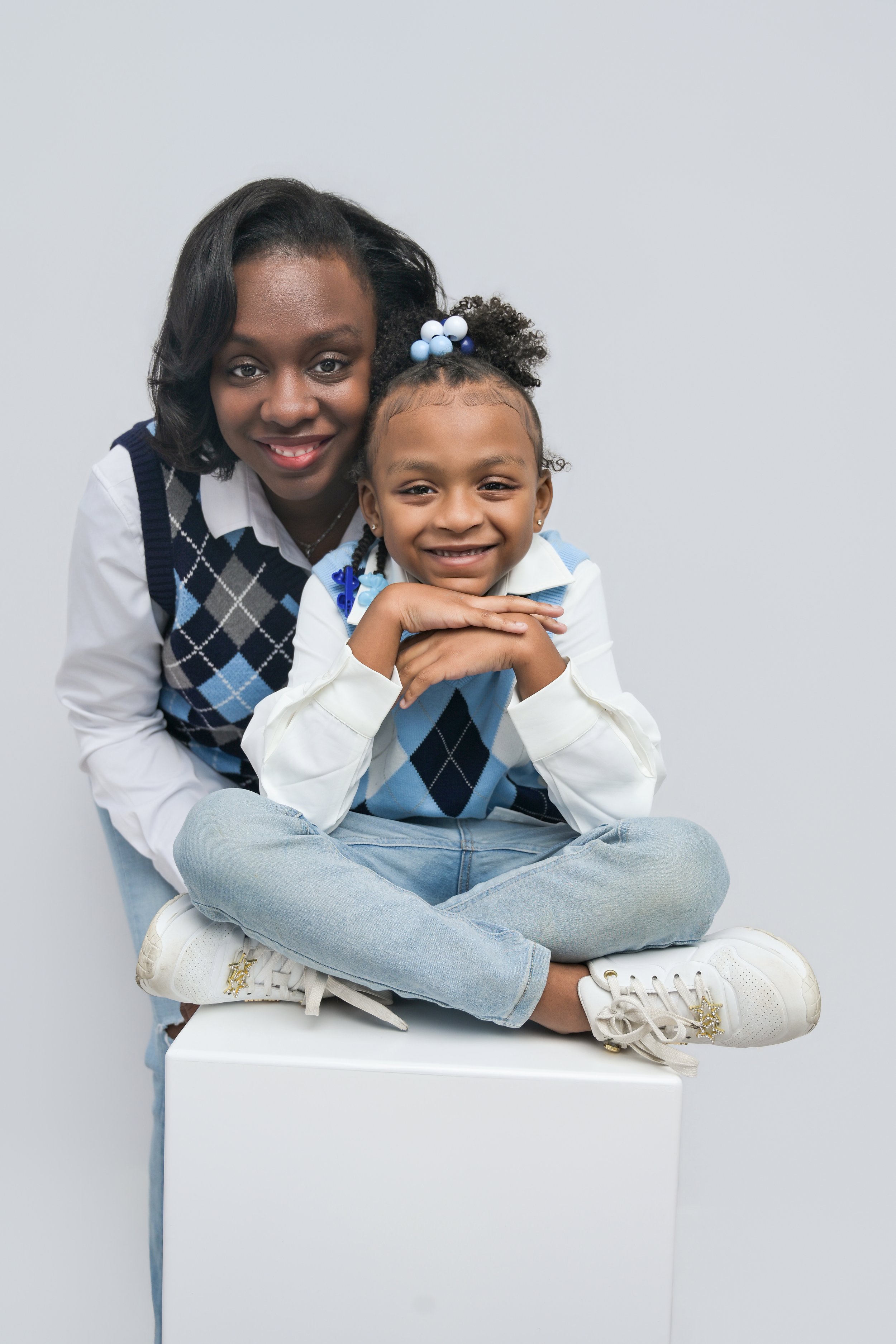 A woman and a young girl sitting on a white block, smiling at the camera, both wearing blue argyle sweaters, with the girl sitting cross-legged and the woman leaning behind her.