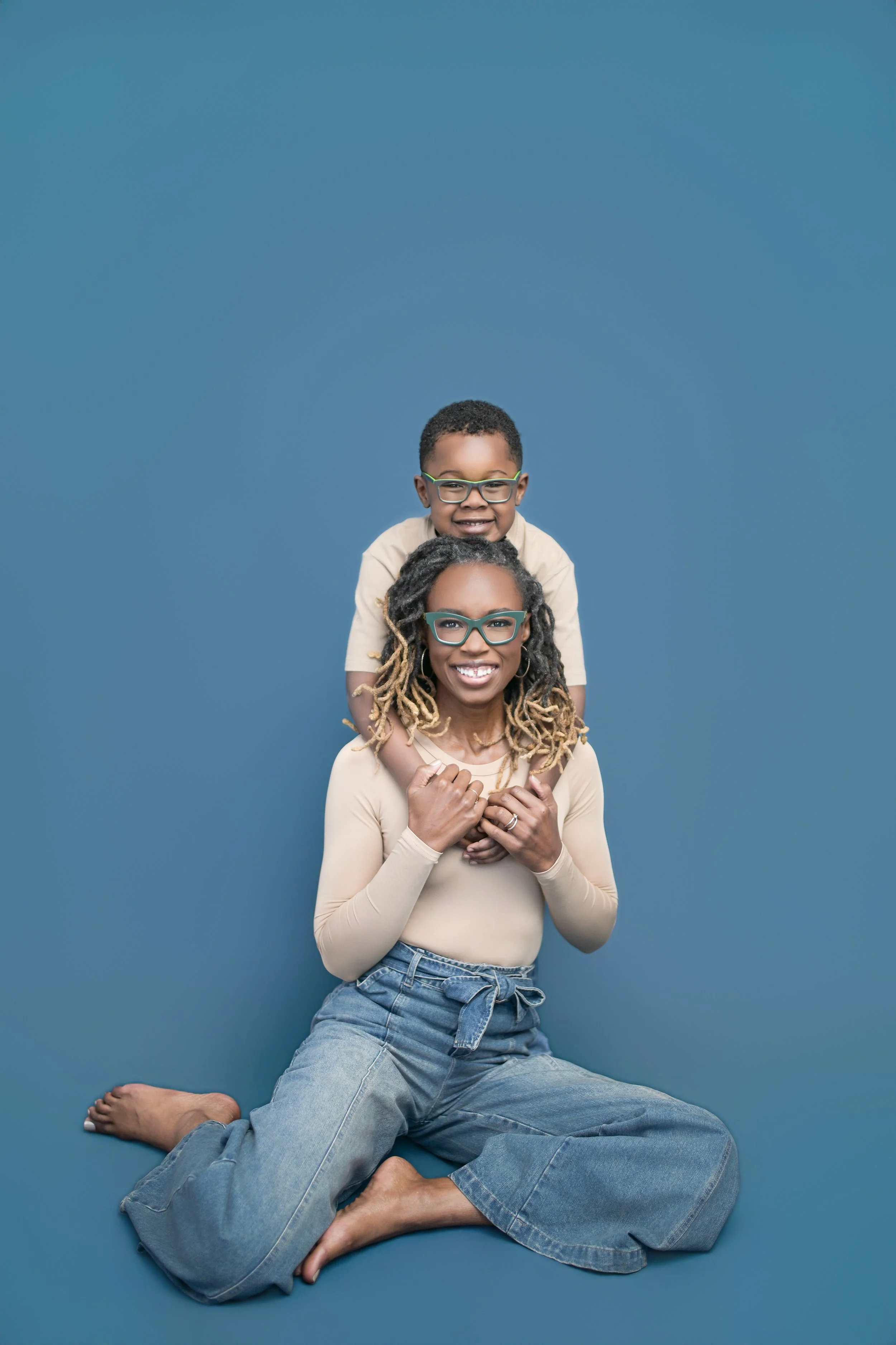 A woman with glasses and long curly hair sitting on the floor with her knees bent and feet flat, smiling at the camera. A young boy with glasses is sitting on her shoulders, also smiling. The background is solid blue. Family portrait.