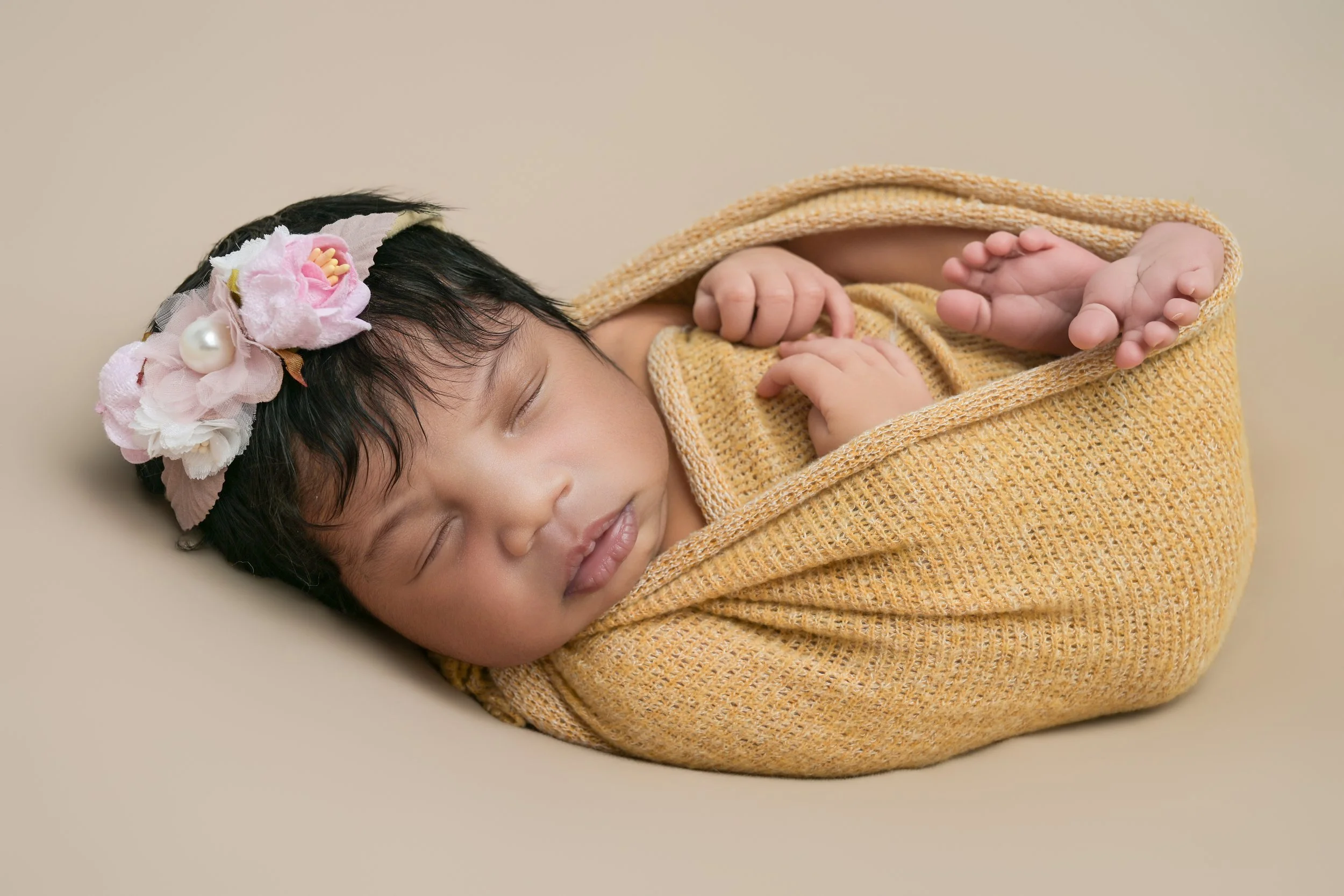 A sleeping baby girl wrapped in a yellow blanket with a floral headband, lying on a beige surface. This is a posed newborn portrait session. 