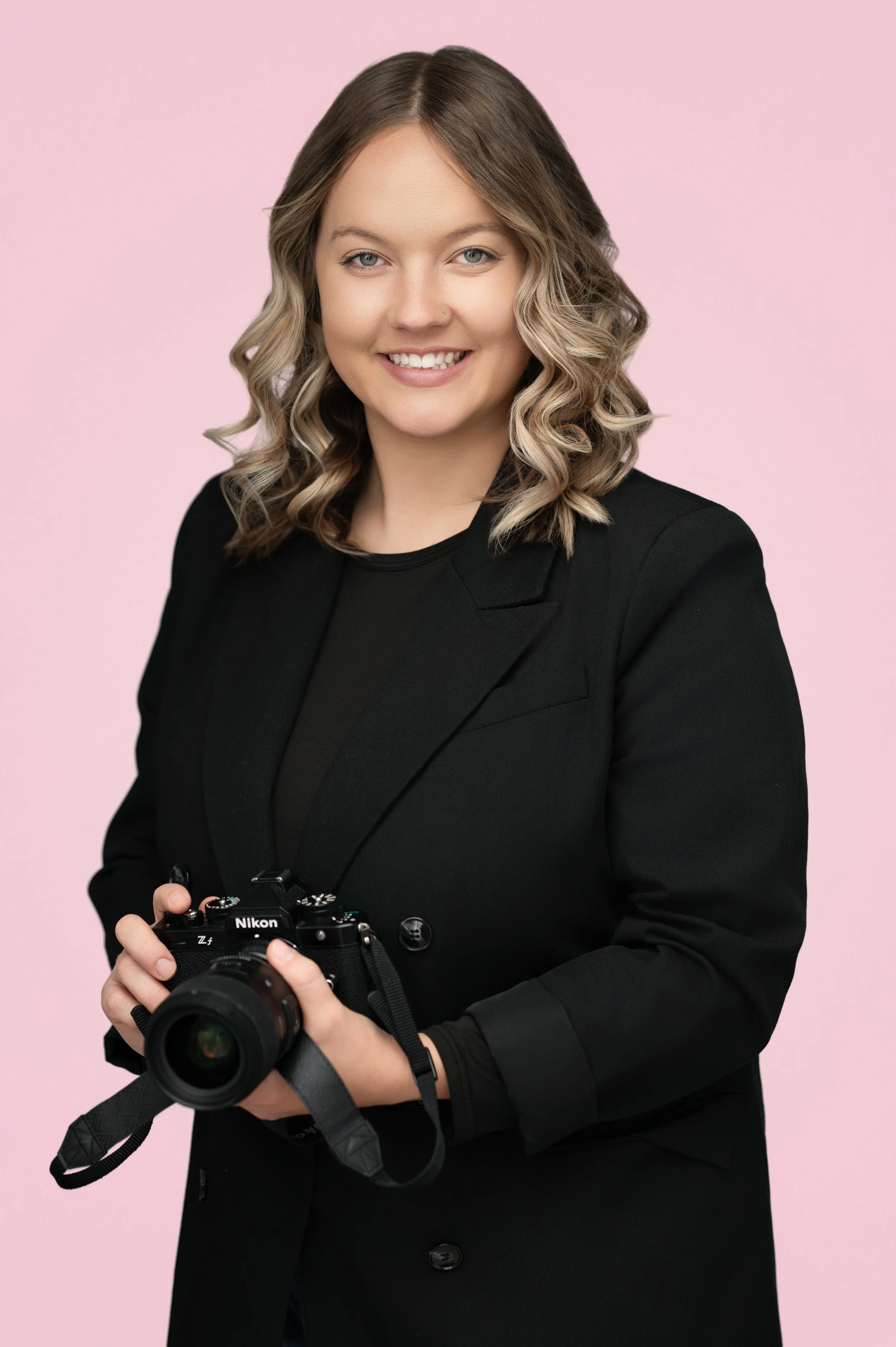 A woman with shoulder-length wavy blonde hair, smiling, wearing a black blazer, holding a Nikon camera, standing against a light pink background. Newborn Portrait Photographer Shelby Wells.