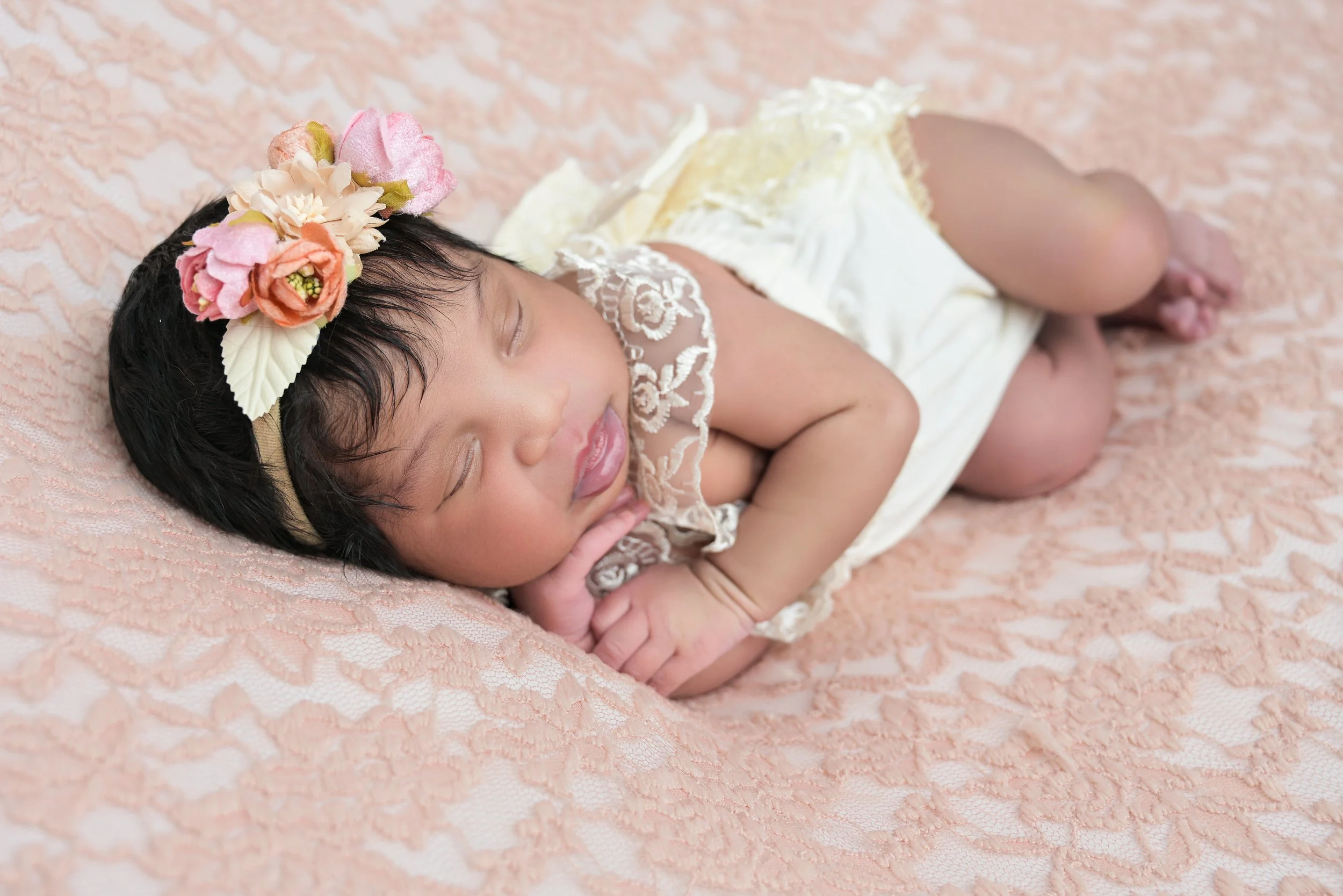 A sleeping baby girl lying on her side on a peach-colored lace blanket, wearing a headband with pink, peach, and cream flowers and a cream dress with lace details. Posed for her newborn portrait session.