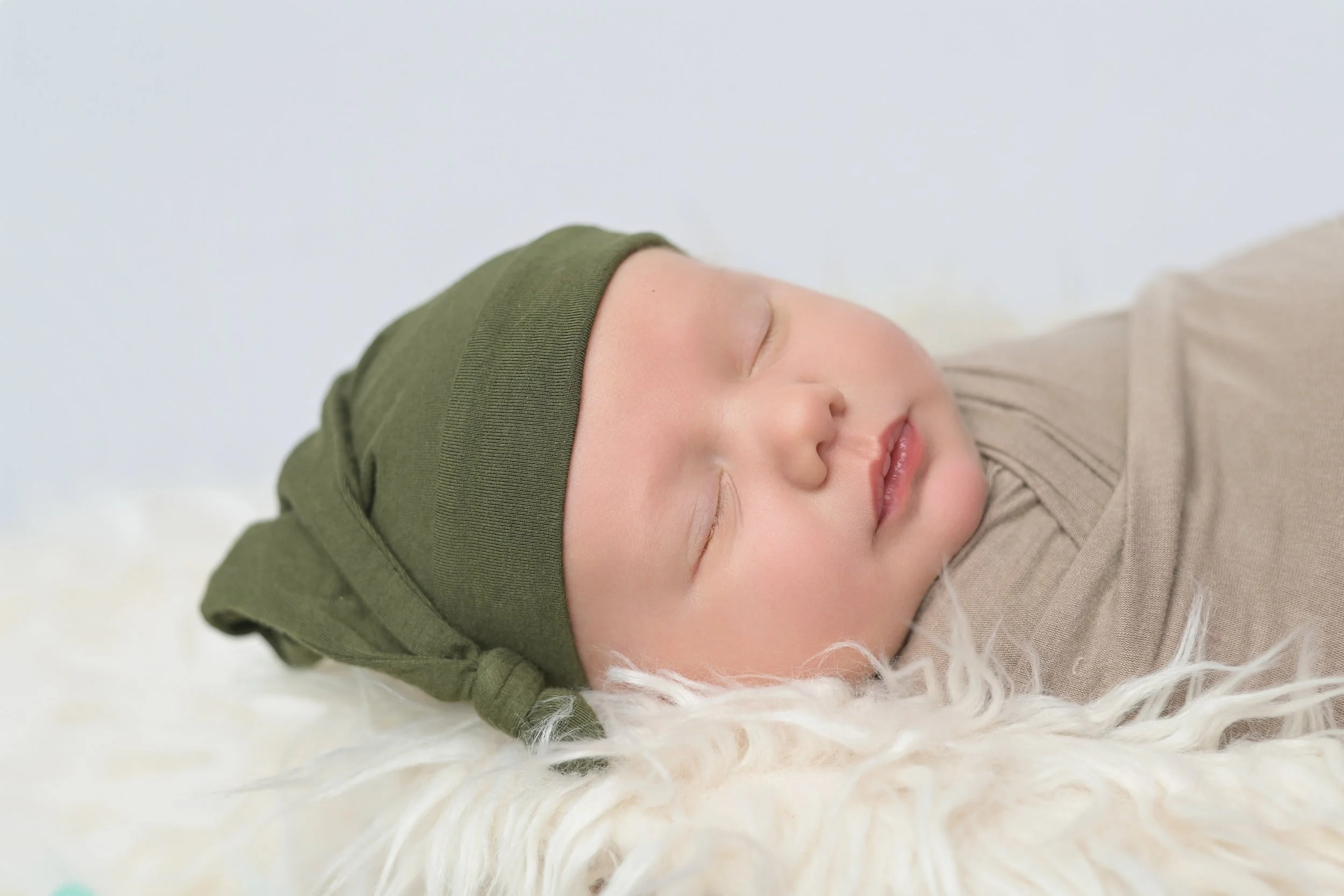 Newborn baby. A sleeping baby in a beige onesie and a green hat, lying on a soft, fluffy white blanket. 