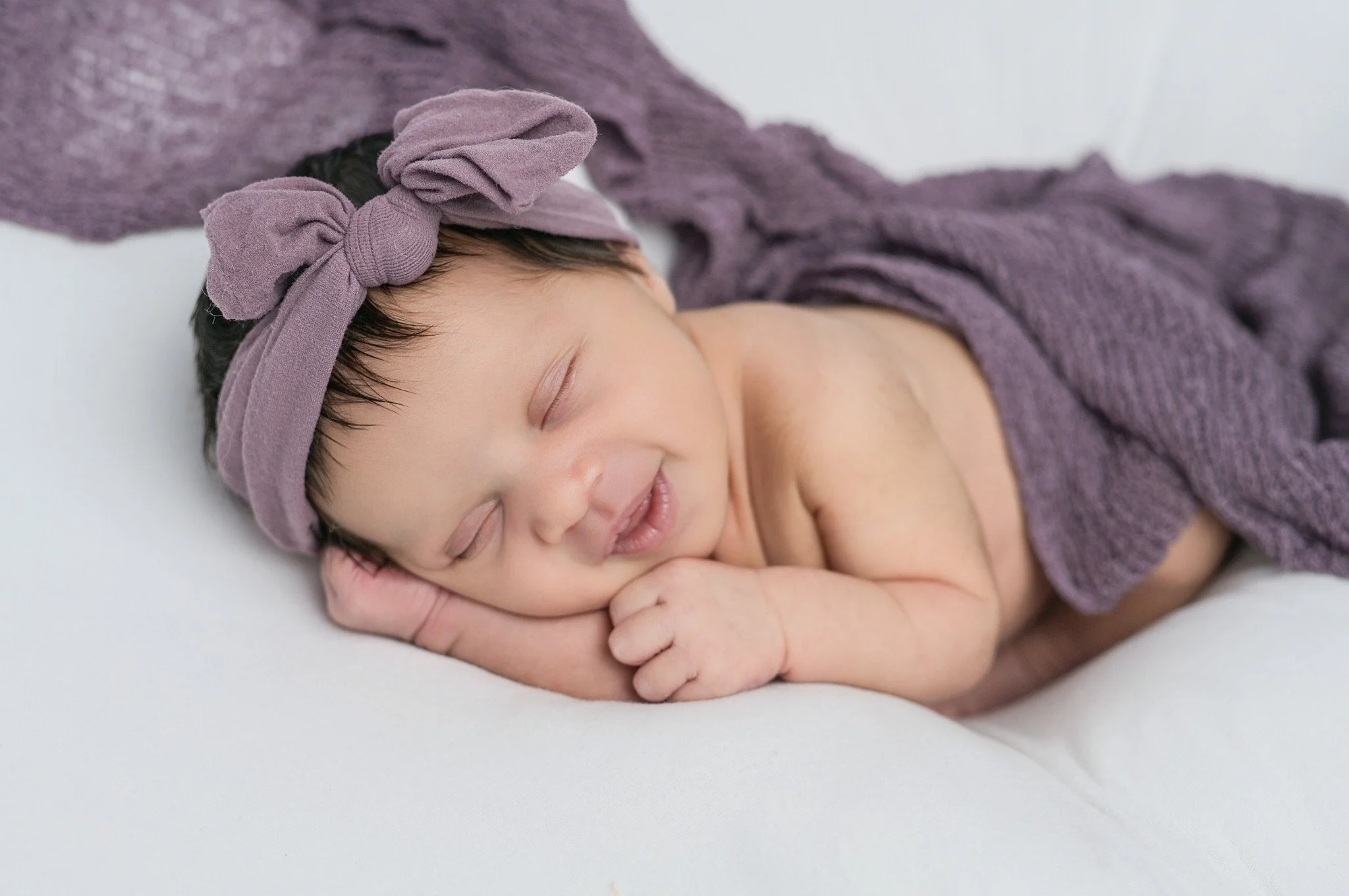 Adorable newborn baby girl sleeping peacefully on a white surface, wearing newborn portrait props, a purple knitted wrap and a matching headband with a bow.