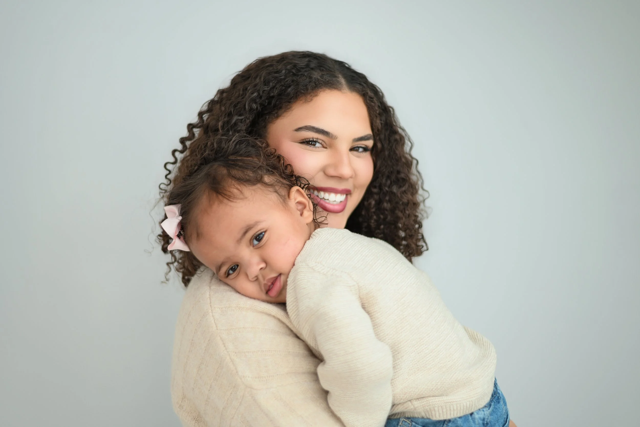 A woman with curly hair smiling and hugging a young girl with a pink bow in her hair, against a plain light gray background. Family Portrait session.