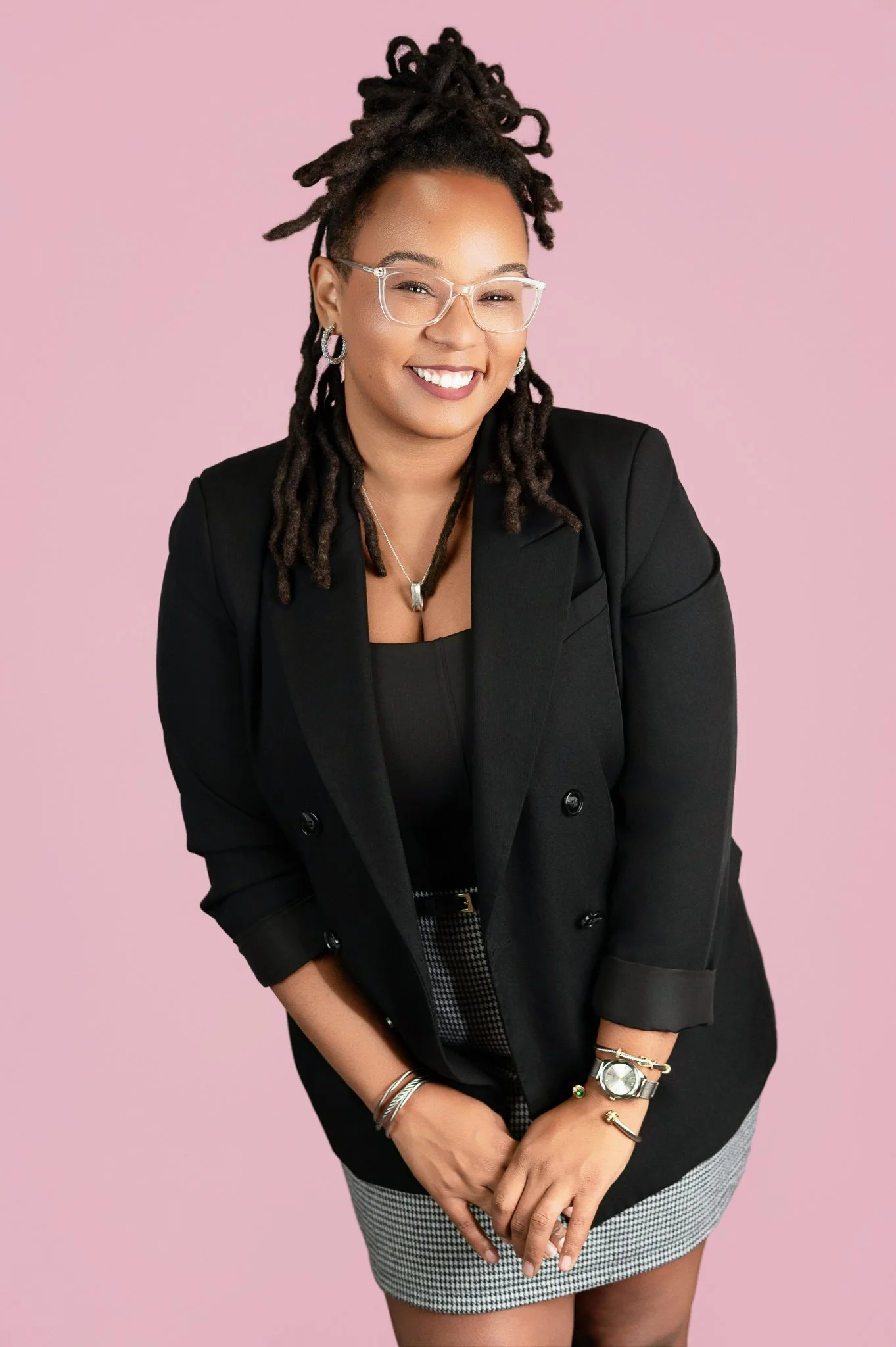A smiling woman with glasses and dreadlocks wearing a black blazer and houndstooth skirt, standing against a pink background.  Black-owned photography studio Chanel N. Wells-Henderson
