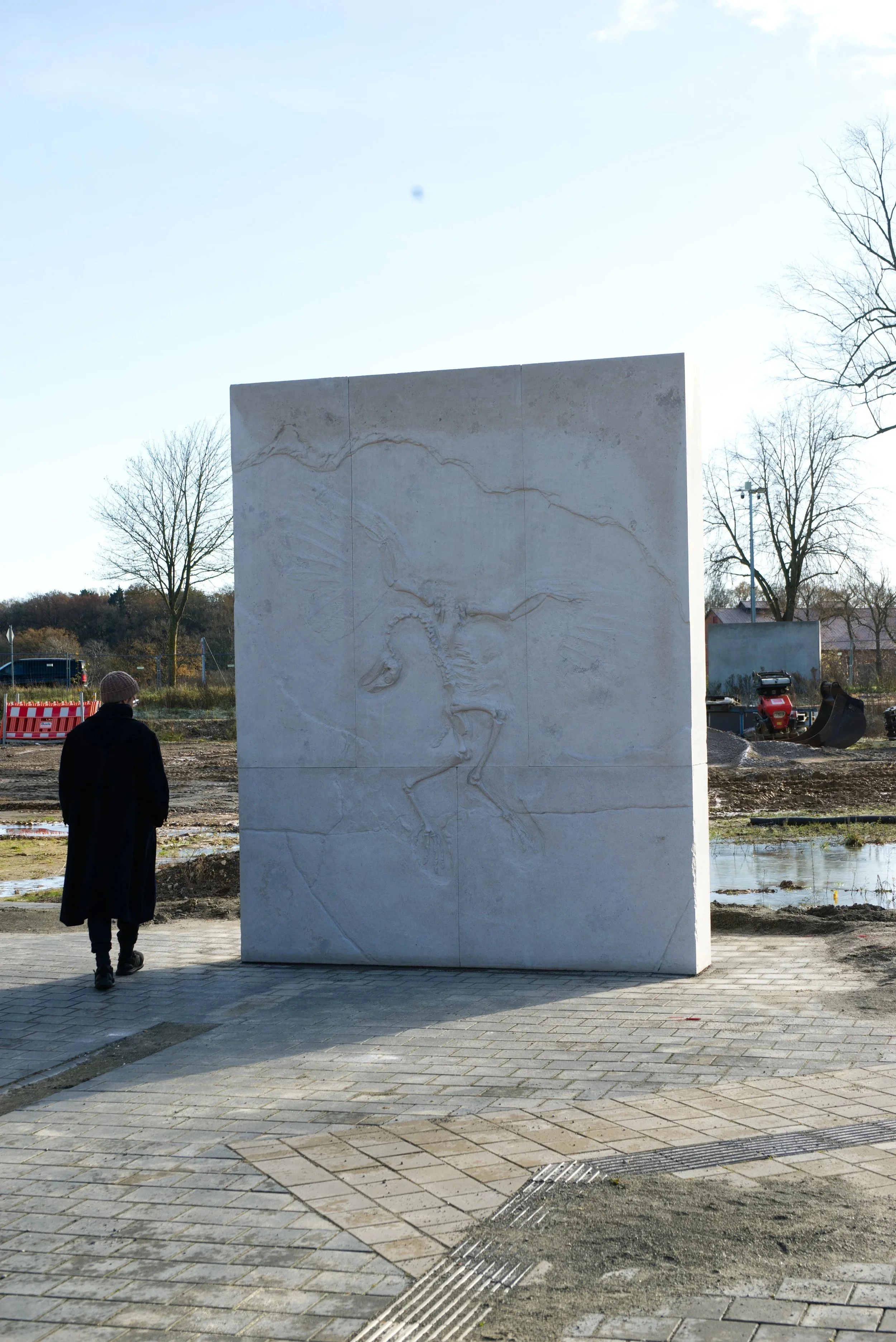 A person dressed in a black coat and a knit cap walks towards a large white stone slab with a carved dinosaur outline on it, outdoors in an area under construction.
