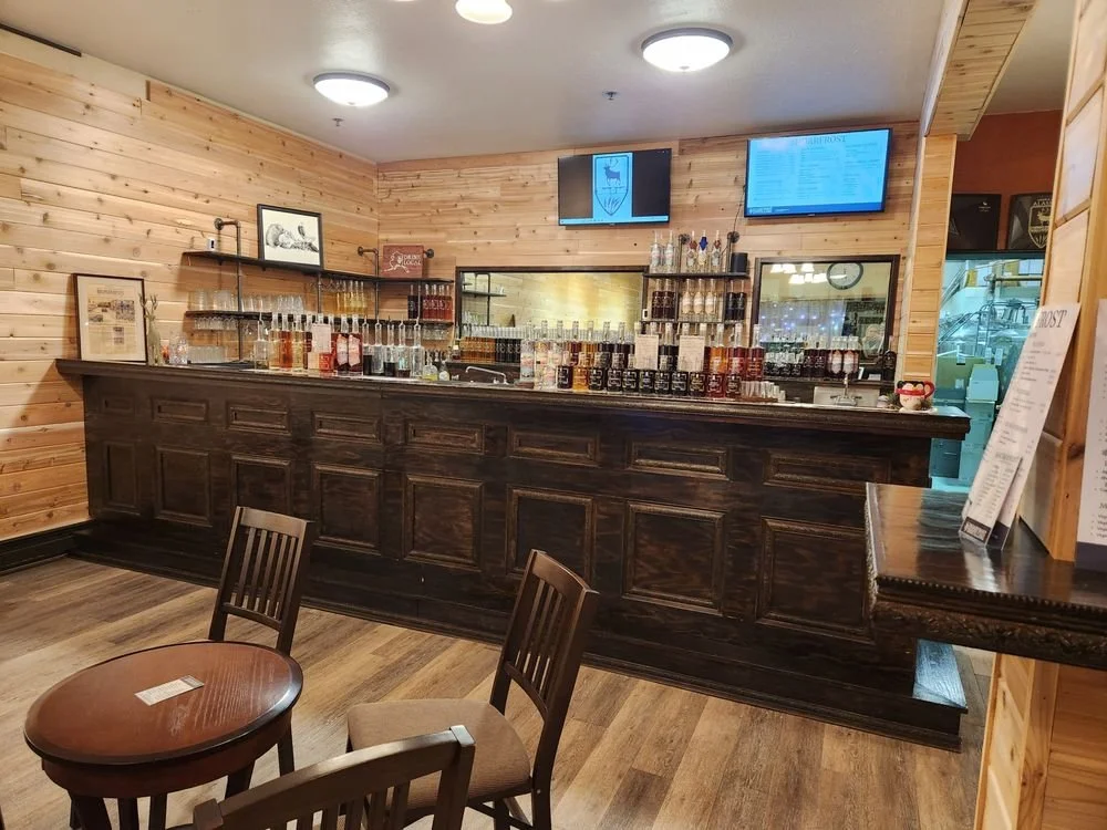 A wooden bar counter with various bottles and glasses behind it, in a cozy restaurant or cafe setting with wooden walls and a small table with chairs in the foreground.