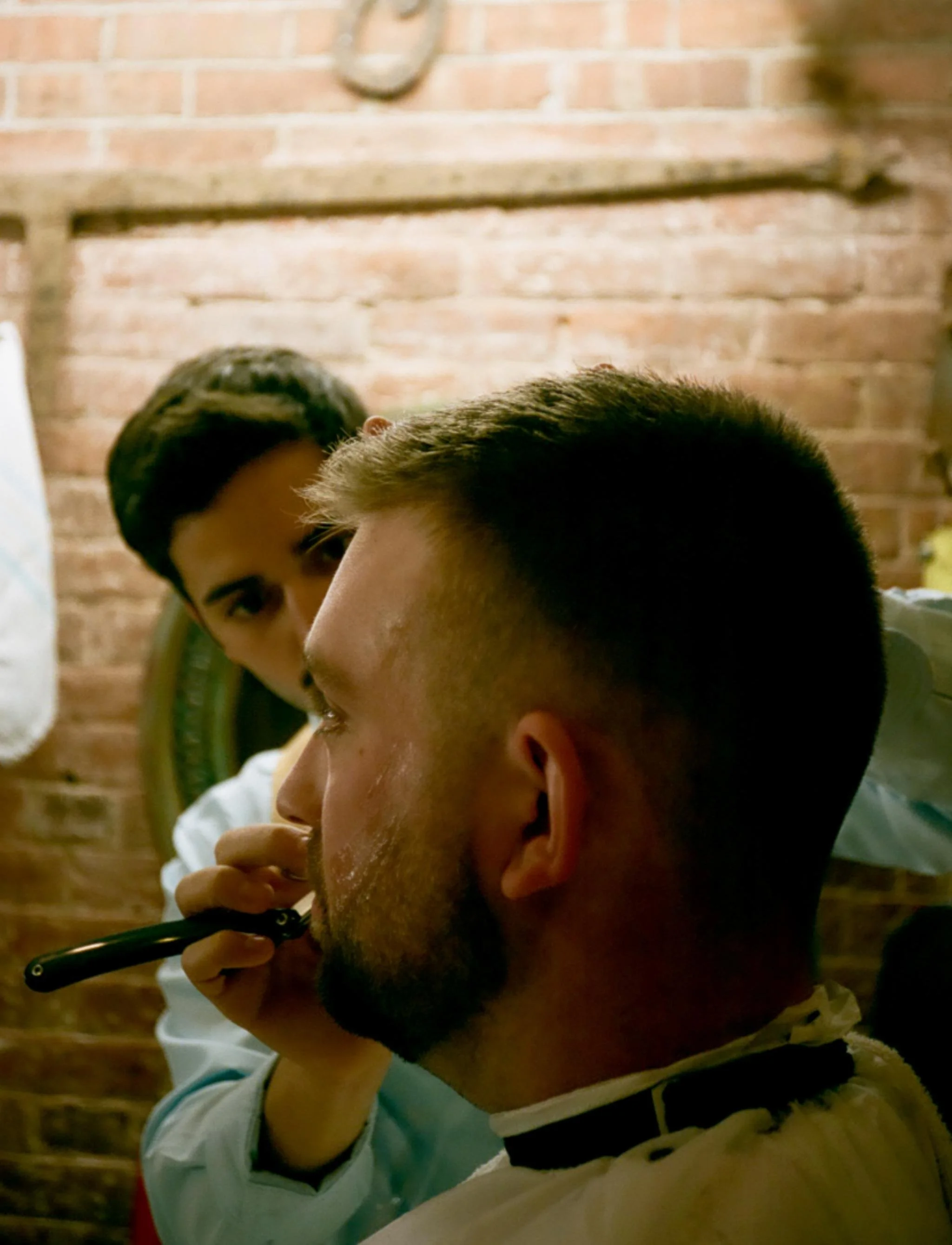 A man getting a shave with a straight razor at a barbershop, with a woman barber in the background.