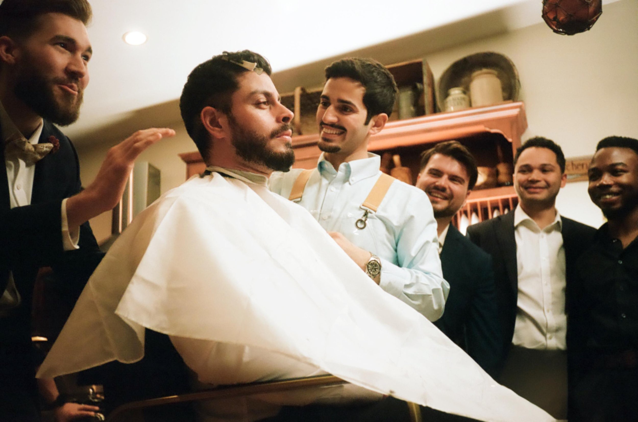 A man receiving a haircut from a barber surrounded by five smiling men in suits in a barbershop.