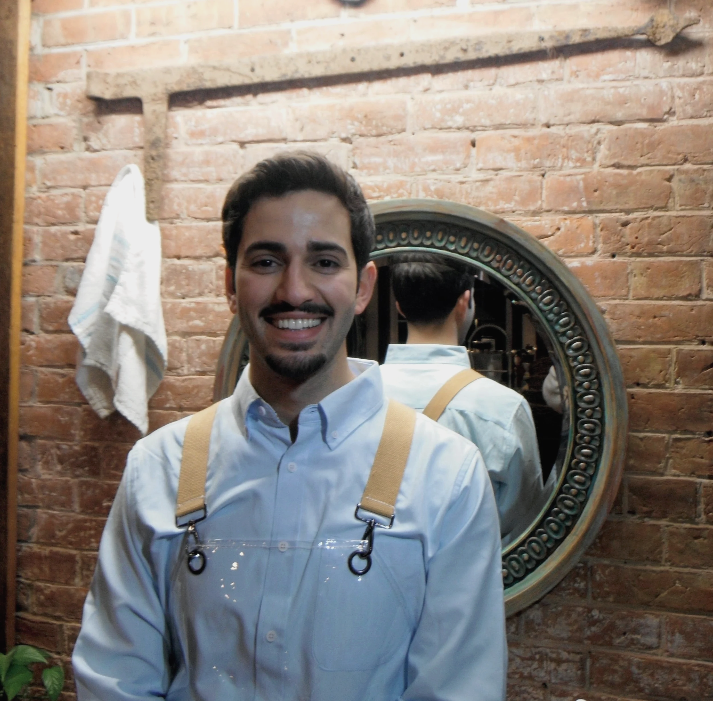Smiling man with dark hair and beard, wearing a white shirt with suspenders, standing in front of a brick wall with a decorative circular mirror and a white towel hanging on the wall.