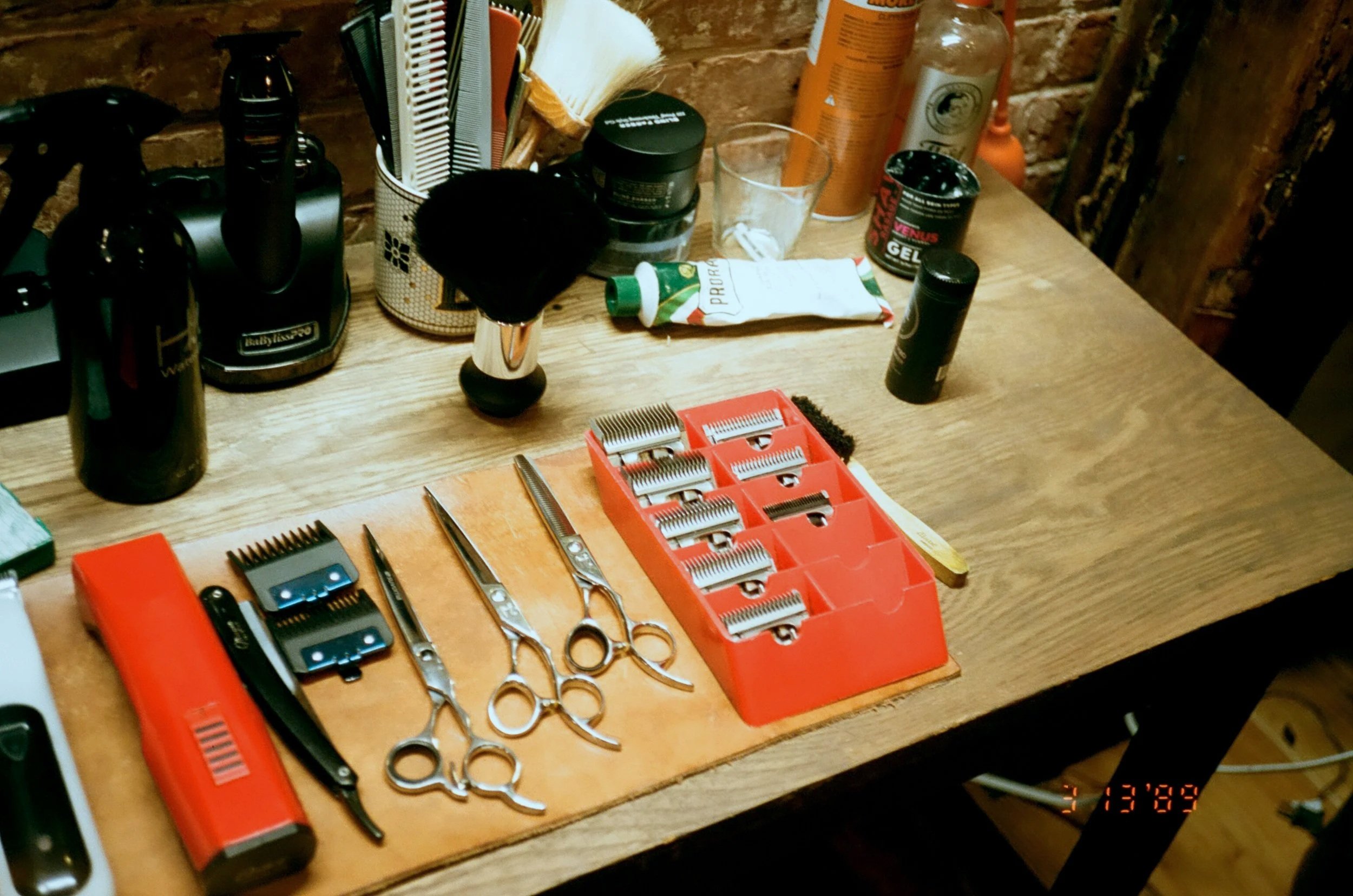 Barber's work station with hair cutting tools including scissors, clippers, a straight razor, and a razor blade holder on a wooden table, with various hair products and brushes in the background.