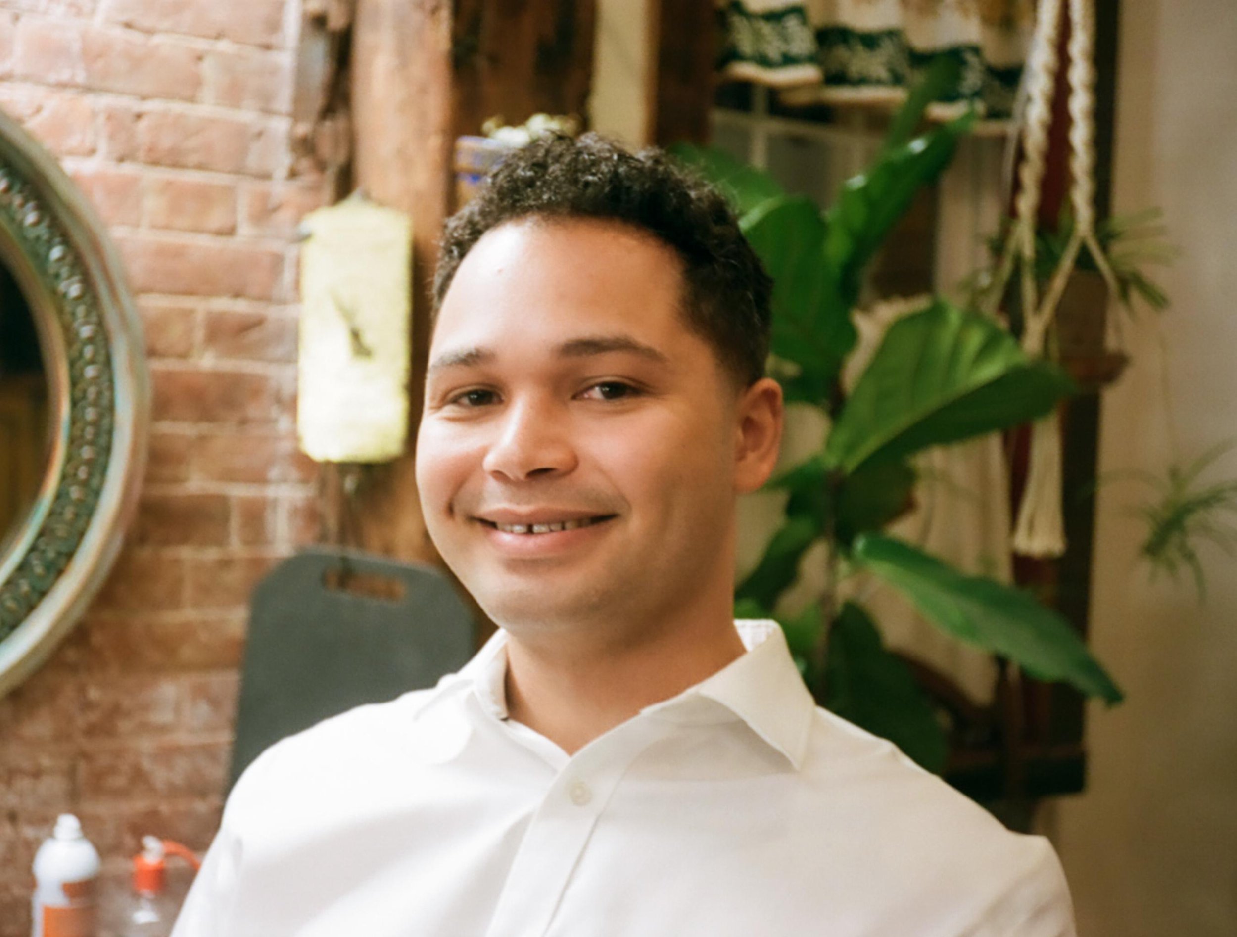Smiling man in a white shirt in a restaurant or cafe with a brick wall, plants, and decorations in the background.