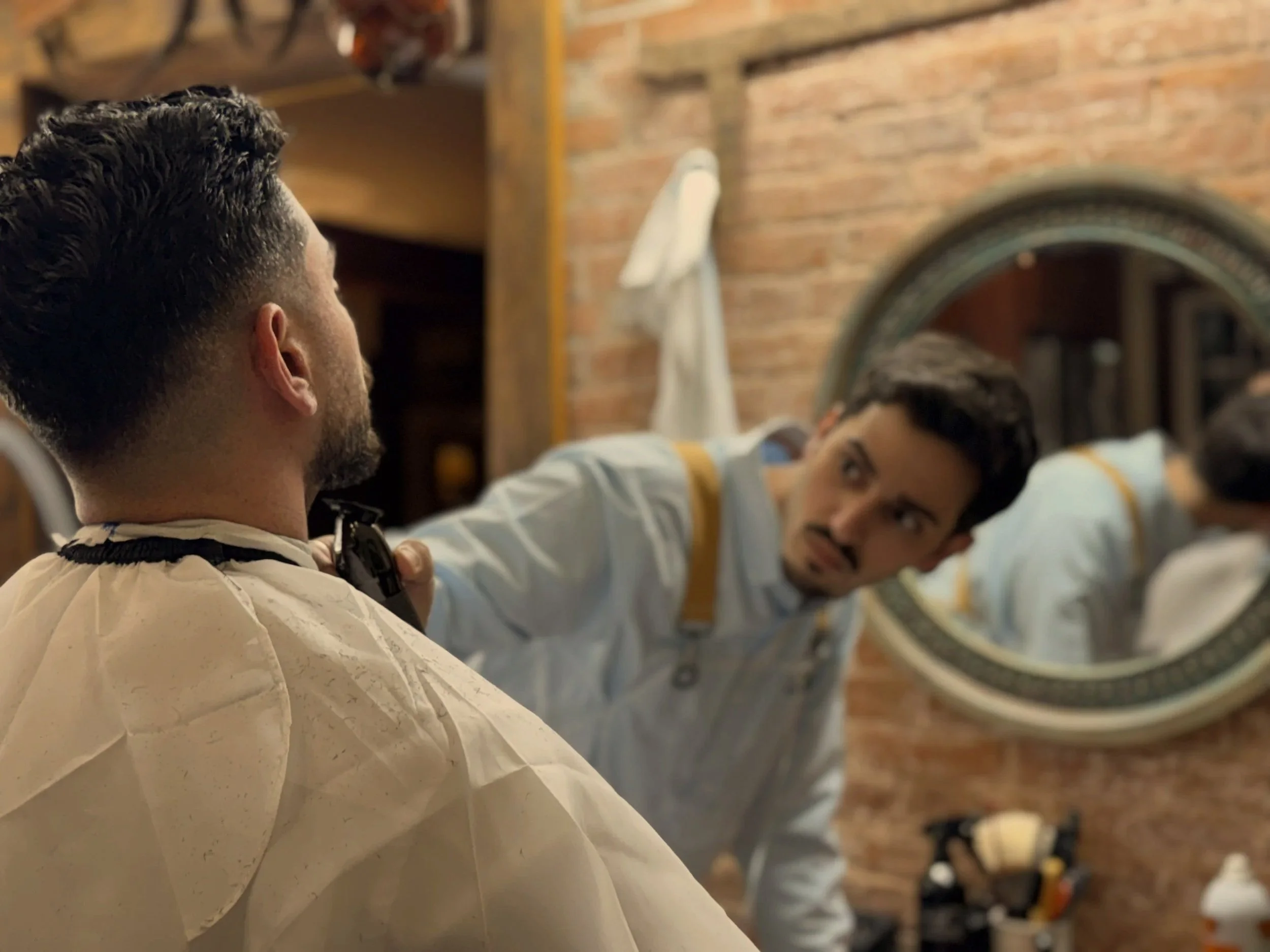 A man getting a haircut at a barbershop, looking into a mirror while a barber trims his hair.