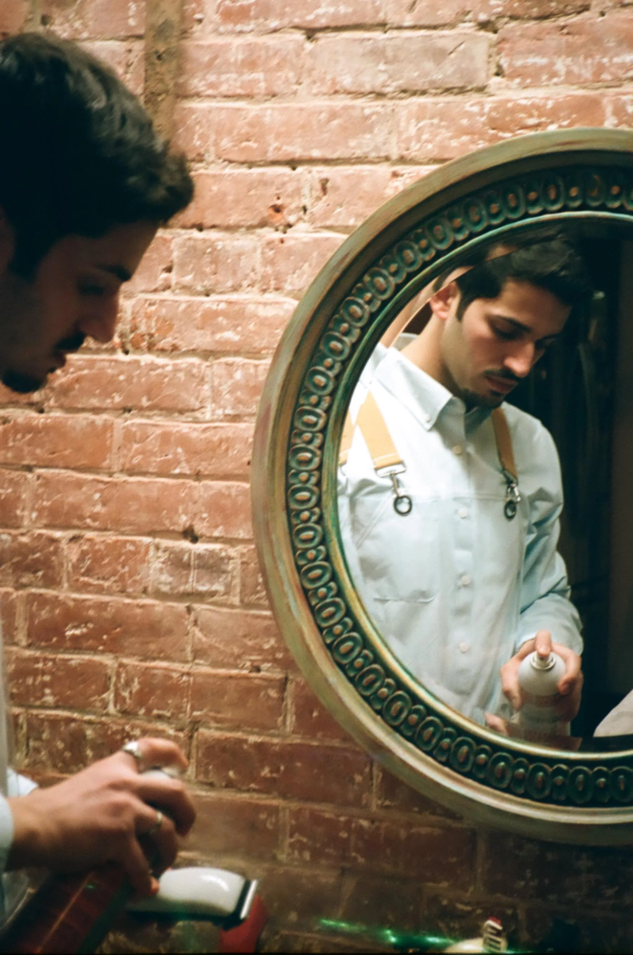 A man looking into a mirror while holding a spray bottle, next to a brick wall.