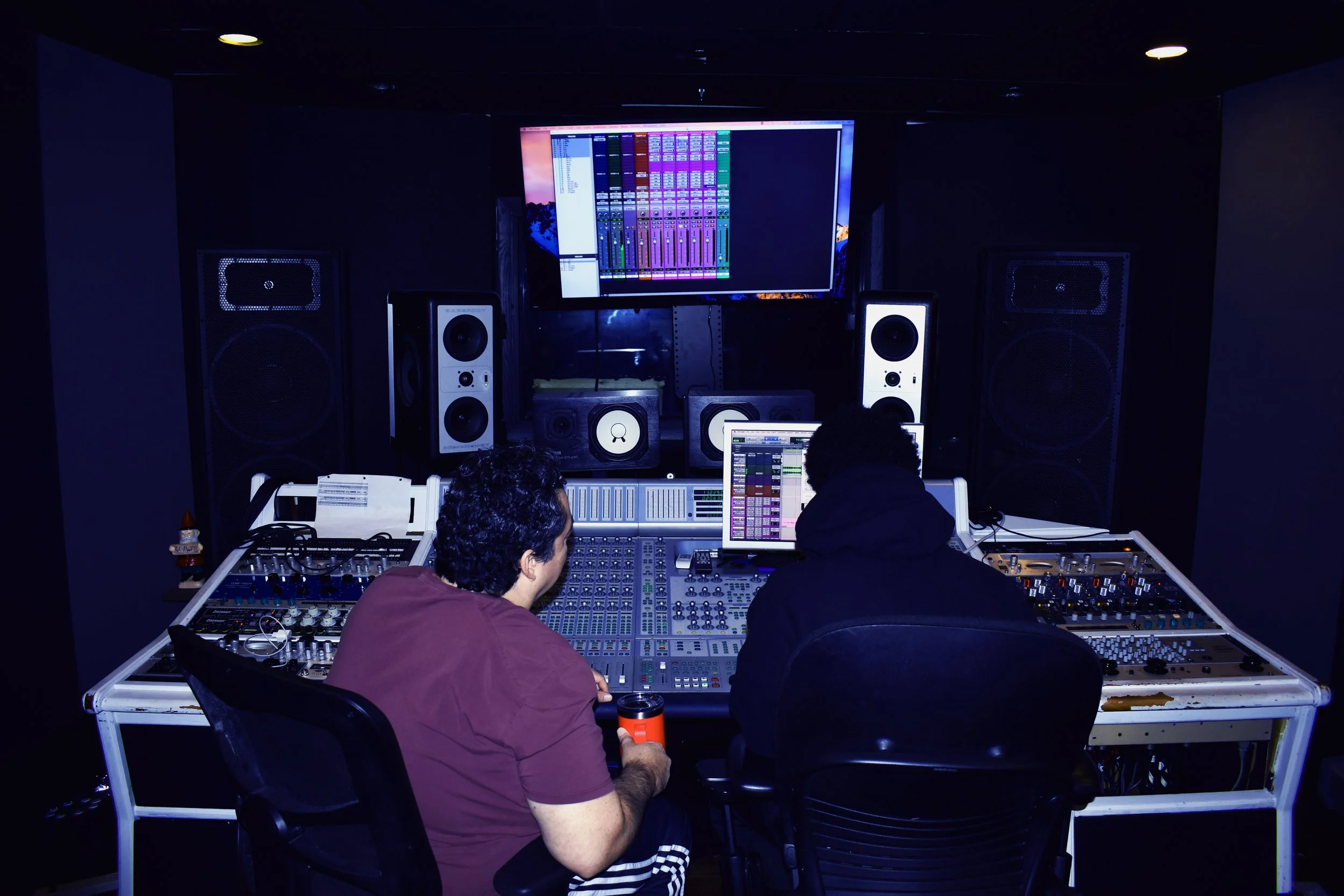Two people working at a professional recording or music mixing studio, with audio equipment and large speakers, facing a computer screen displaying music software.