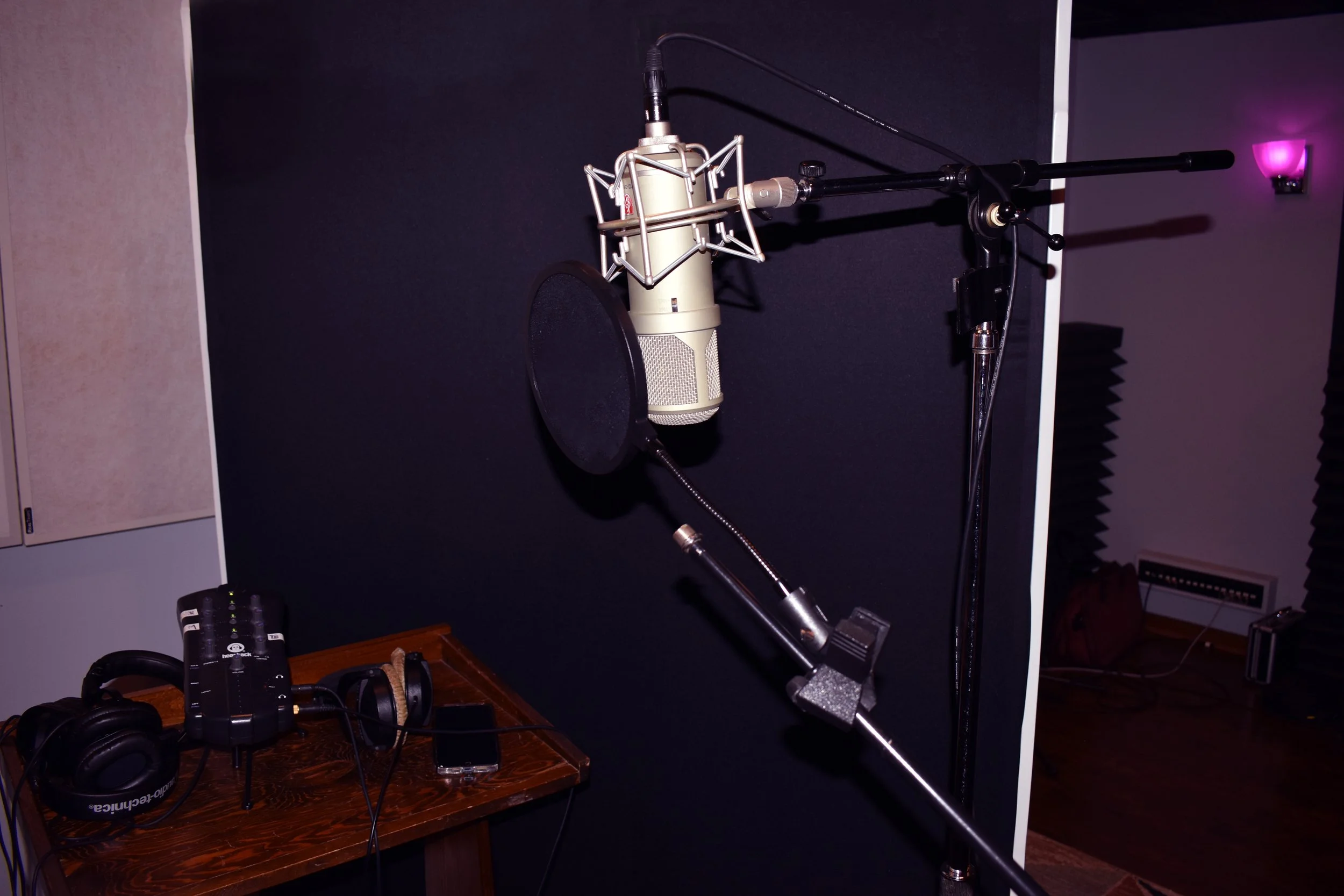 Recording studio corner with a microphone on a stand, a pop filter, a mixing console, headphones, and a smartphone on a wooden table, with soundproofing panels on the wall.