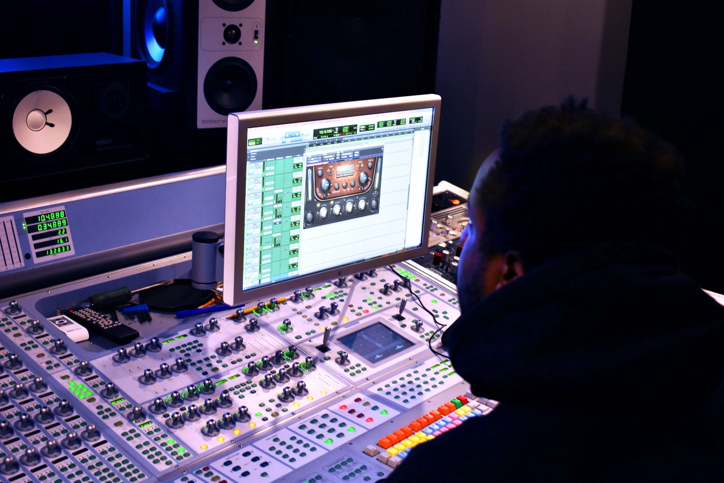 A person working on a music mixing console with a computer monitor displaying audio editing software, in a studio with speakers and audio equipment.
