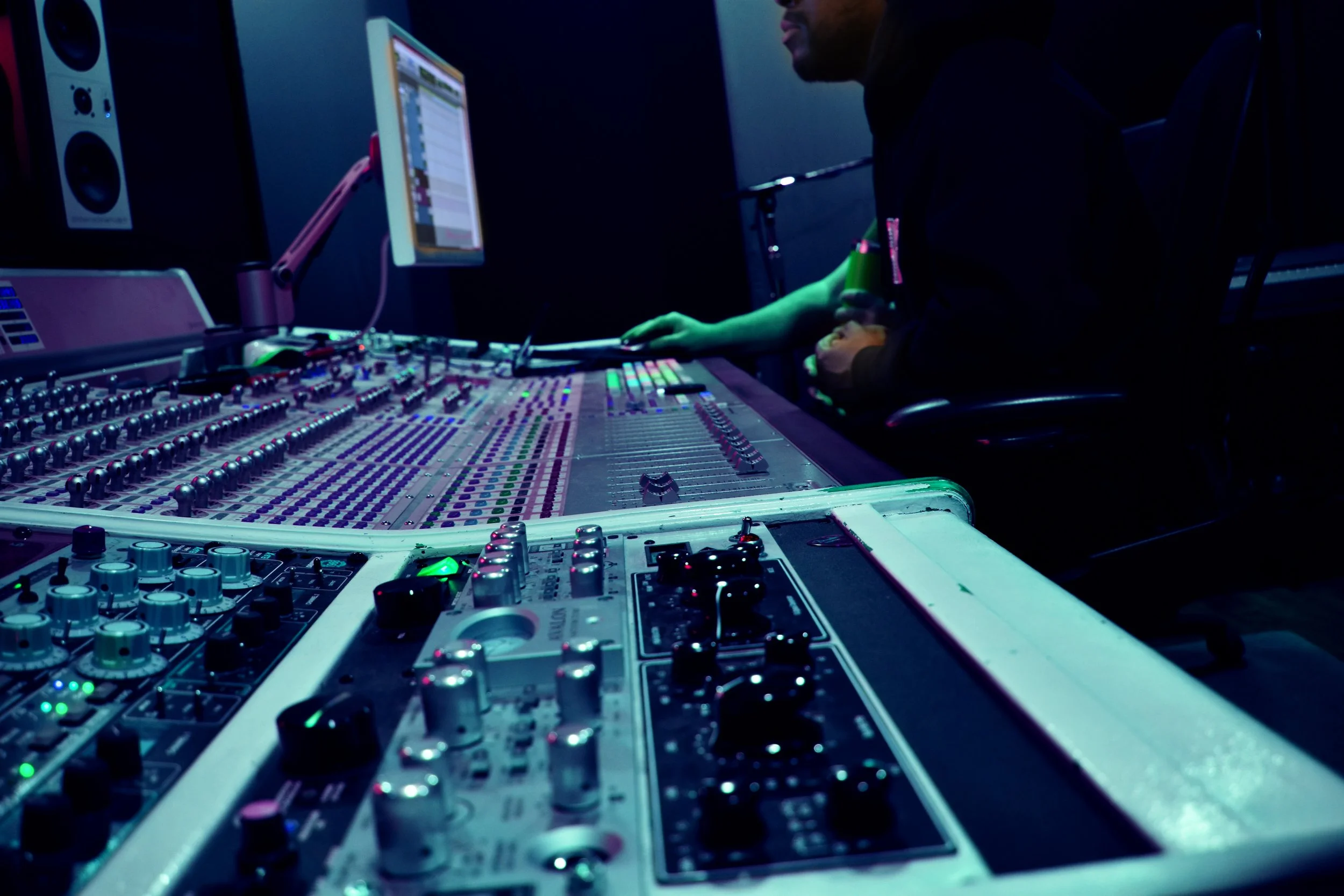 Close-up of audio mixing console with various knobs and sliders in a dimly lit studio, with a person working on a computer monitor in the background.