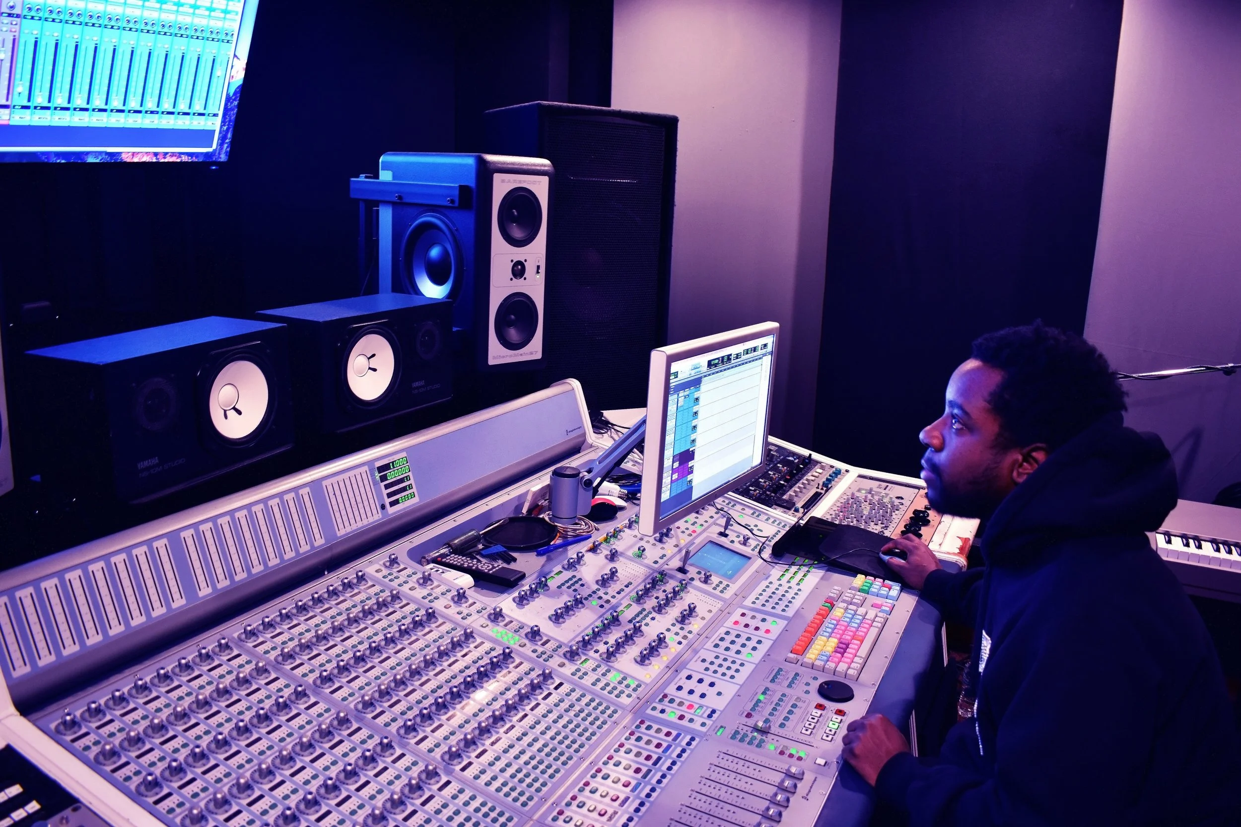 Alontae operating a mixing console and music production equipment in a recording studio, with multiple monitors, speakers, and audio gear visible.
