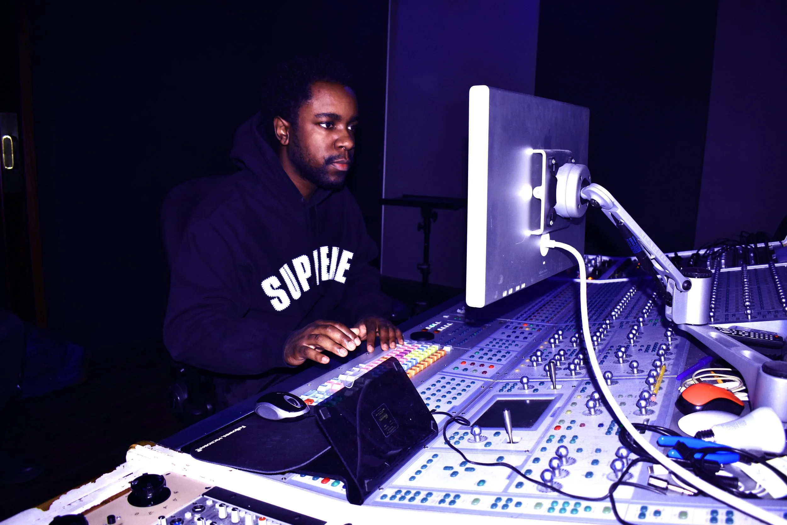 Alontae, wearing a black hoodie with white text, working at a mixing console with a computer monitor in a dark room.