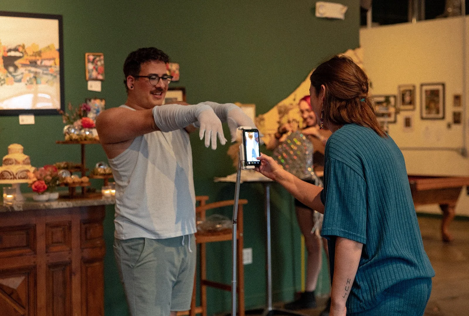 A person in a blue shirt is taking a photo of a man with glasses and a mustache, wearing gloves, in an art gallery or cafe with green walls and artwork on the walls.