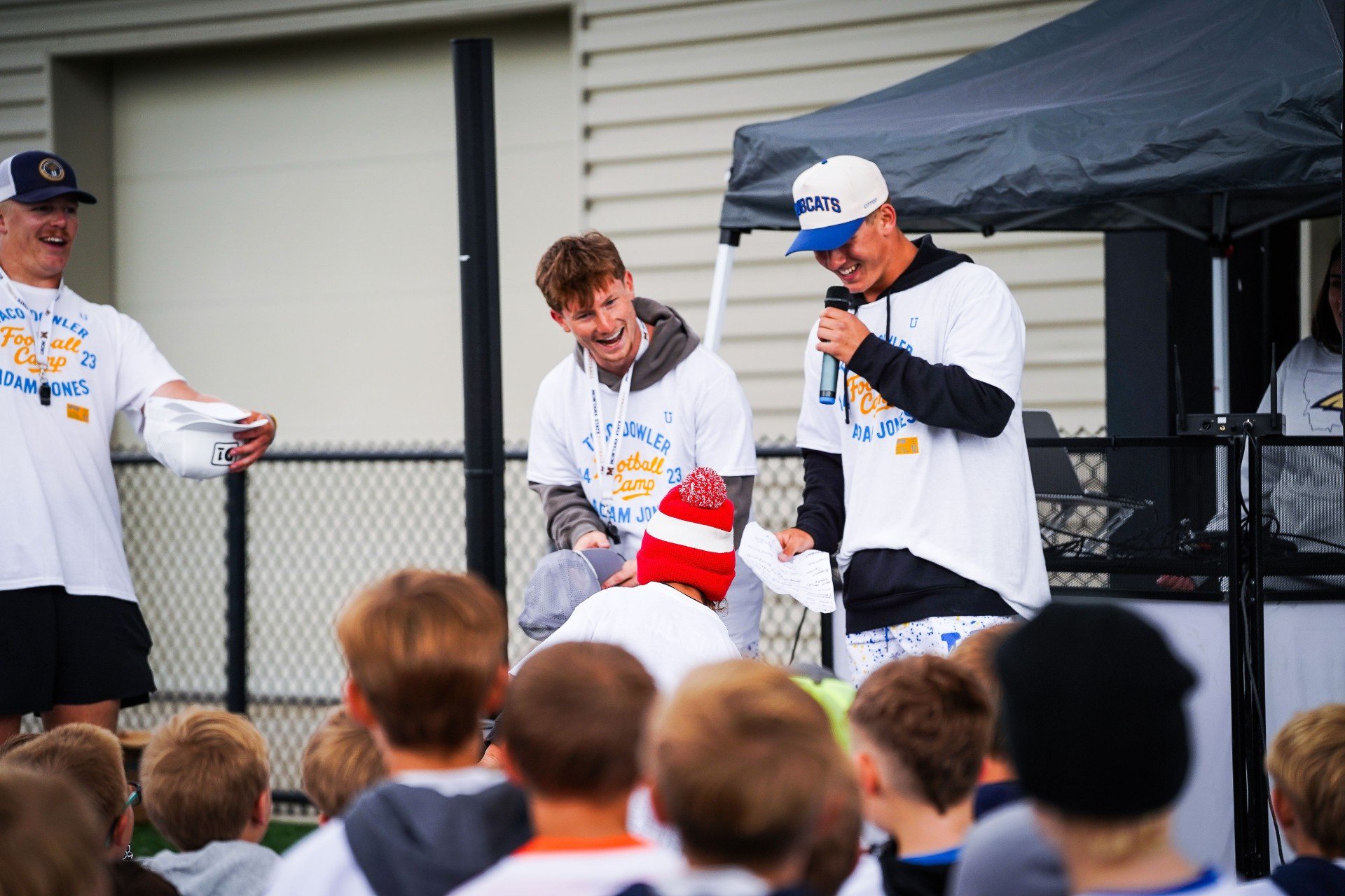 A group of young men at a football camp, some holding papers, laughing and talking on a stage outdoors with children in the audience. One man is holding a microphone and wearing a baseball cap, and everyone is dressed in camp T-shirts.
