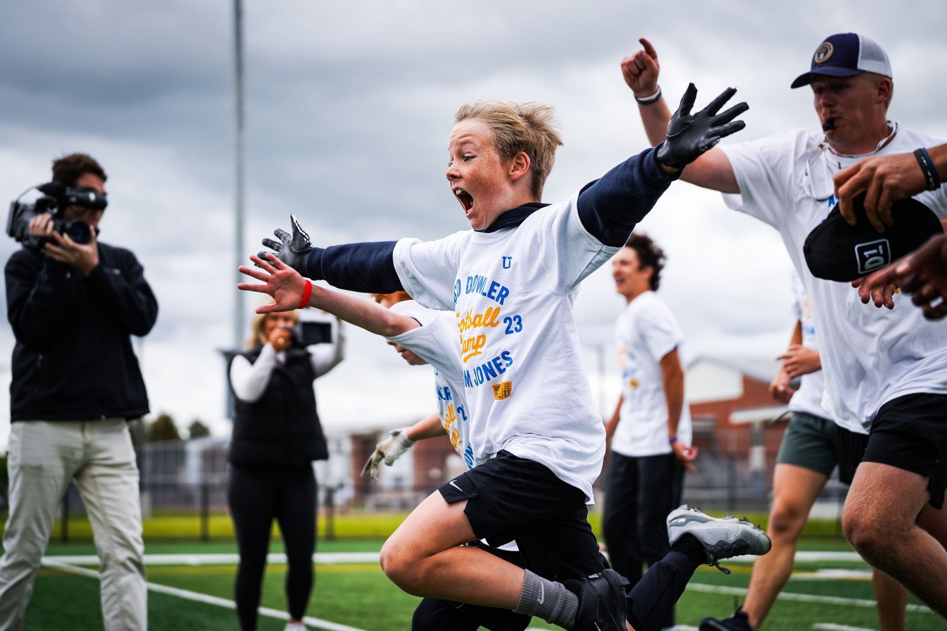 Young person celebrating with arms outstretched on a football field, wearing a white t-shirt with blue and yellow text, surrounded by others and photographers, on an overcast day.