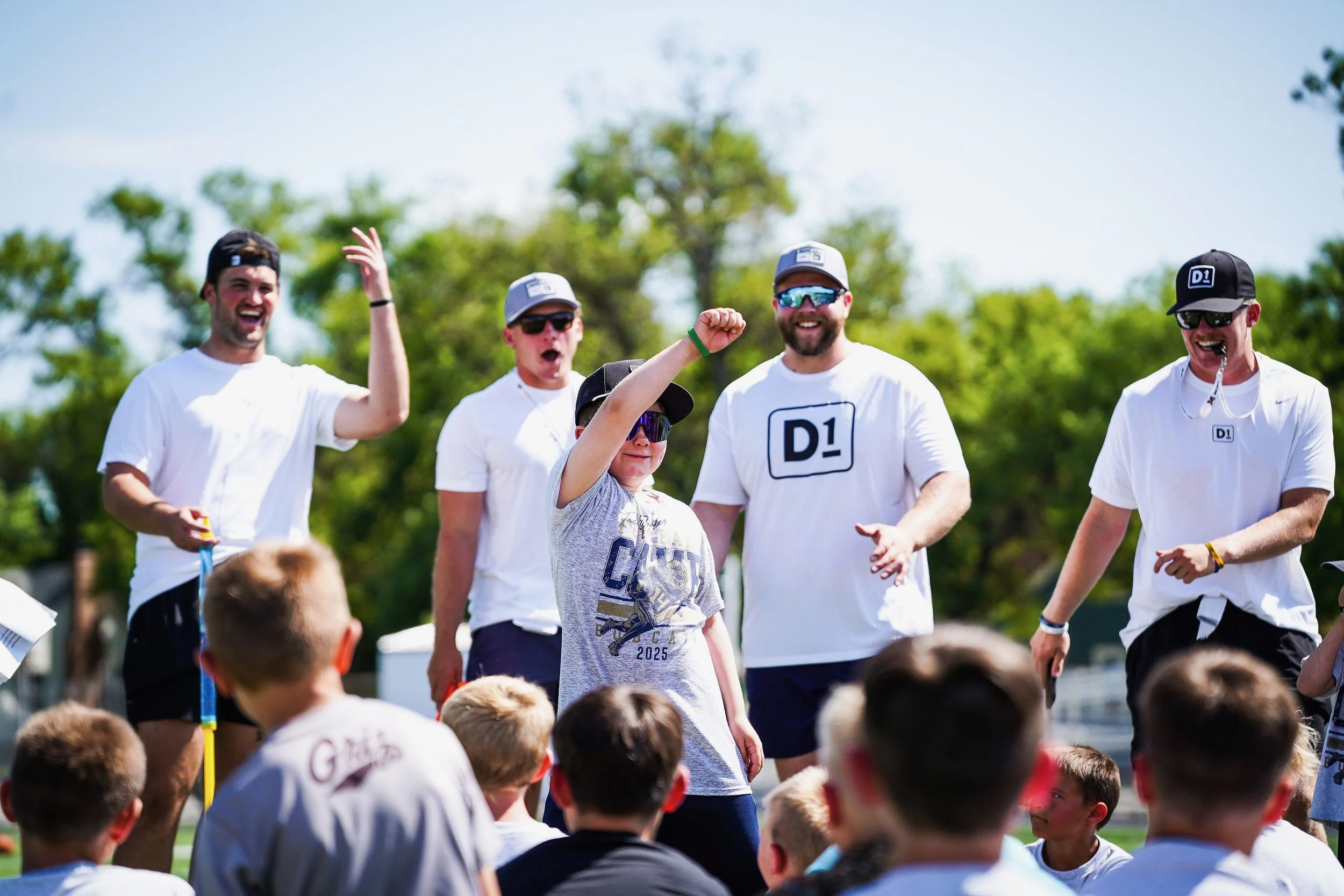 Group of young men and children outdoors on a sunny day, some with sunglasses, celebrating and smiling, with trees in the background.