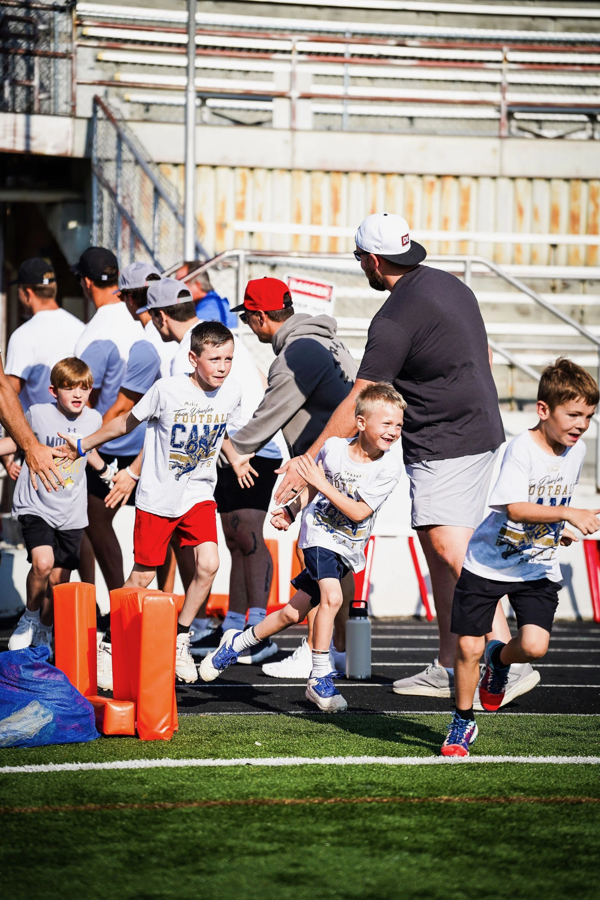 Children participating in a football training session on a field, running and high-fiving each other with coaches in the background.