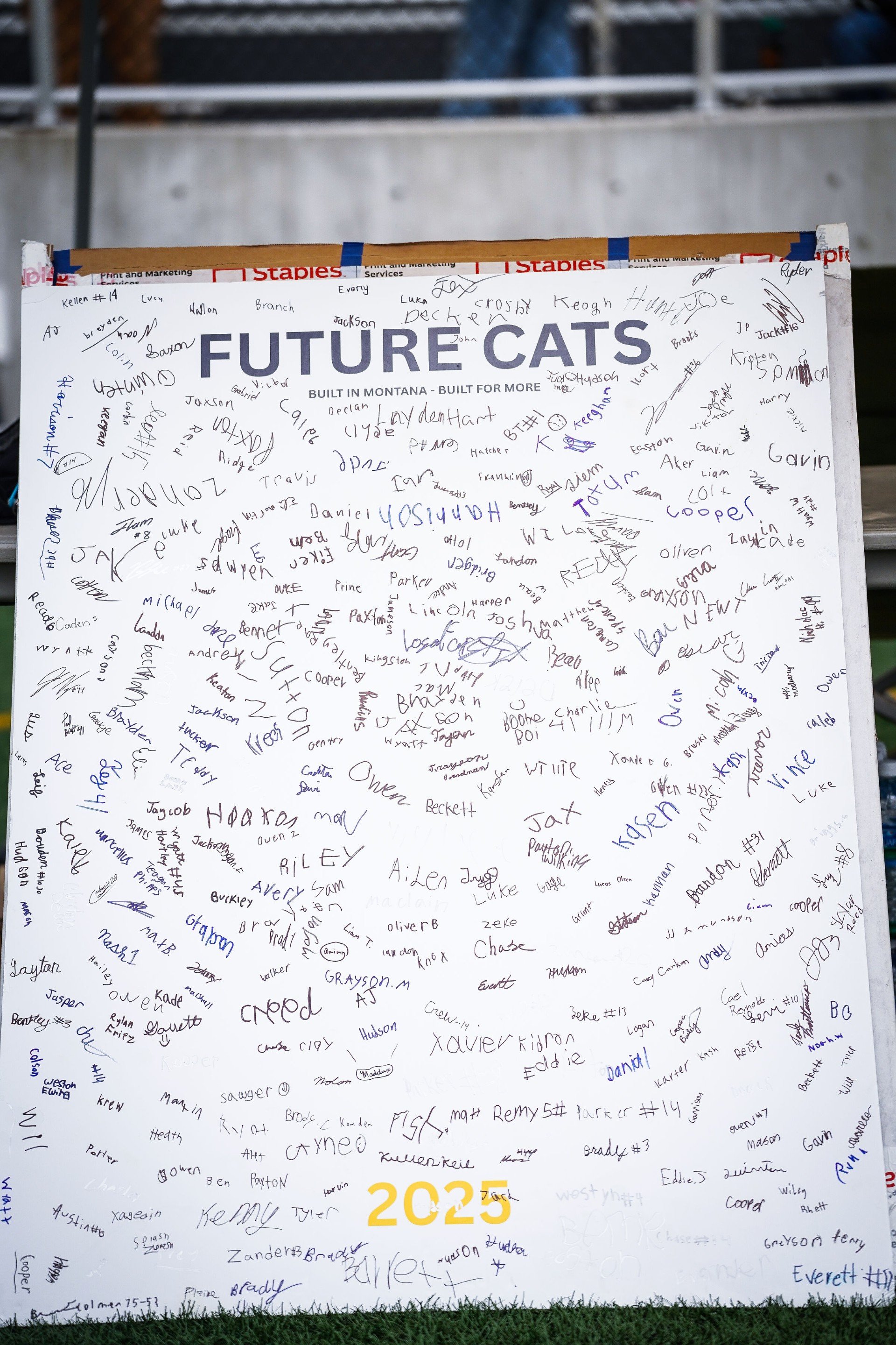 A large white poster board with various signatures and names, with the words "FUTURE CATS" and "BUILT IN MONTANA - BUILT FOR MORE" at the top, and "2023" in yellow at the bottom, situated outdoors on a grassy area.