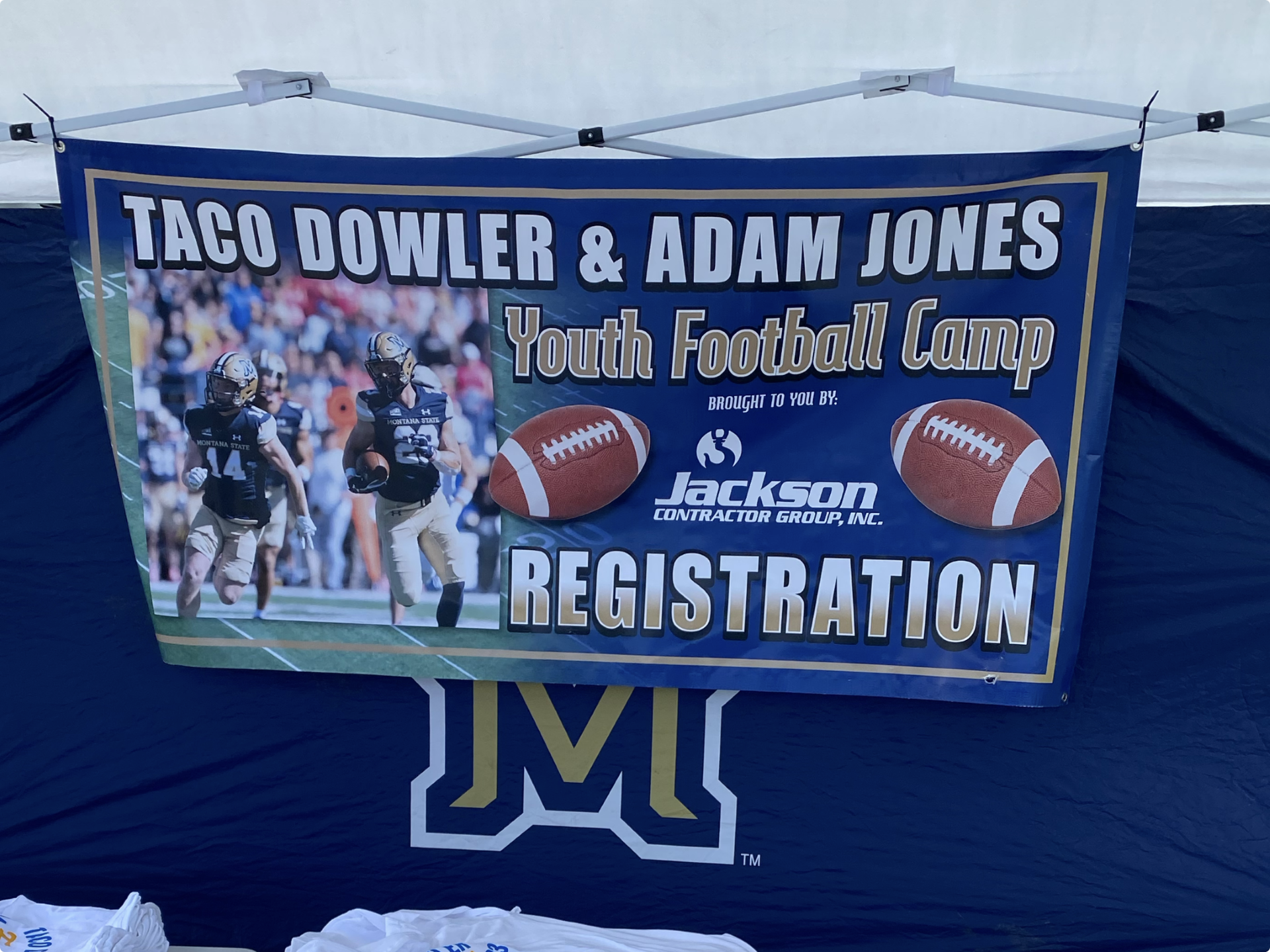 Banner promoting Taco Dowler and Adam Jones Youth Football Camp, featuring a photo of youth football players in jerseys with helmets, two footballs, and the Jackson Contractor Group Inc. logo.