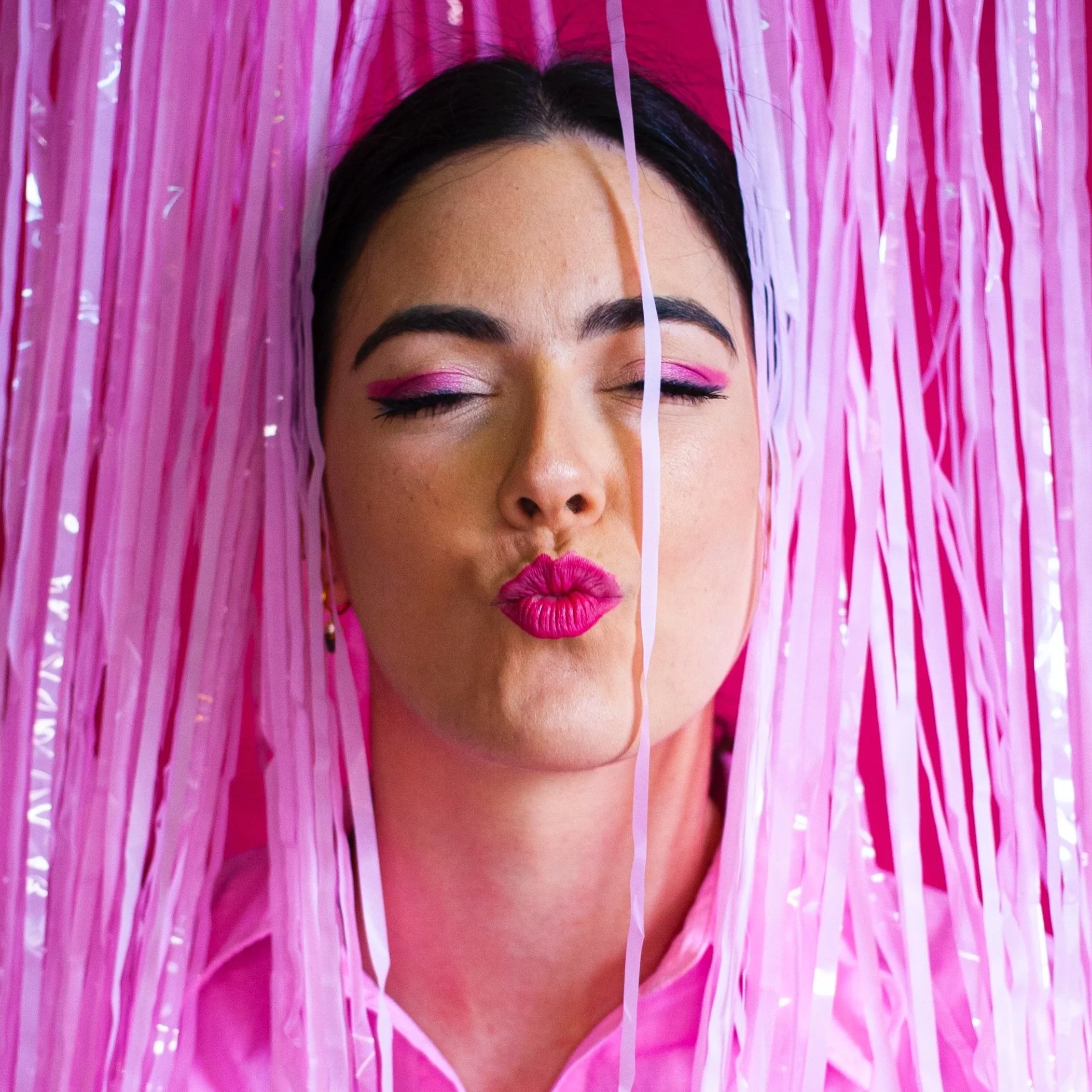 Retrato de una mujer con maquillaje rosado, con labios en forma de beso y ojos cerrados, rodeada de decoraciones rosas. Contenido de redes sociales.
