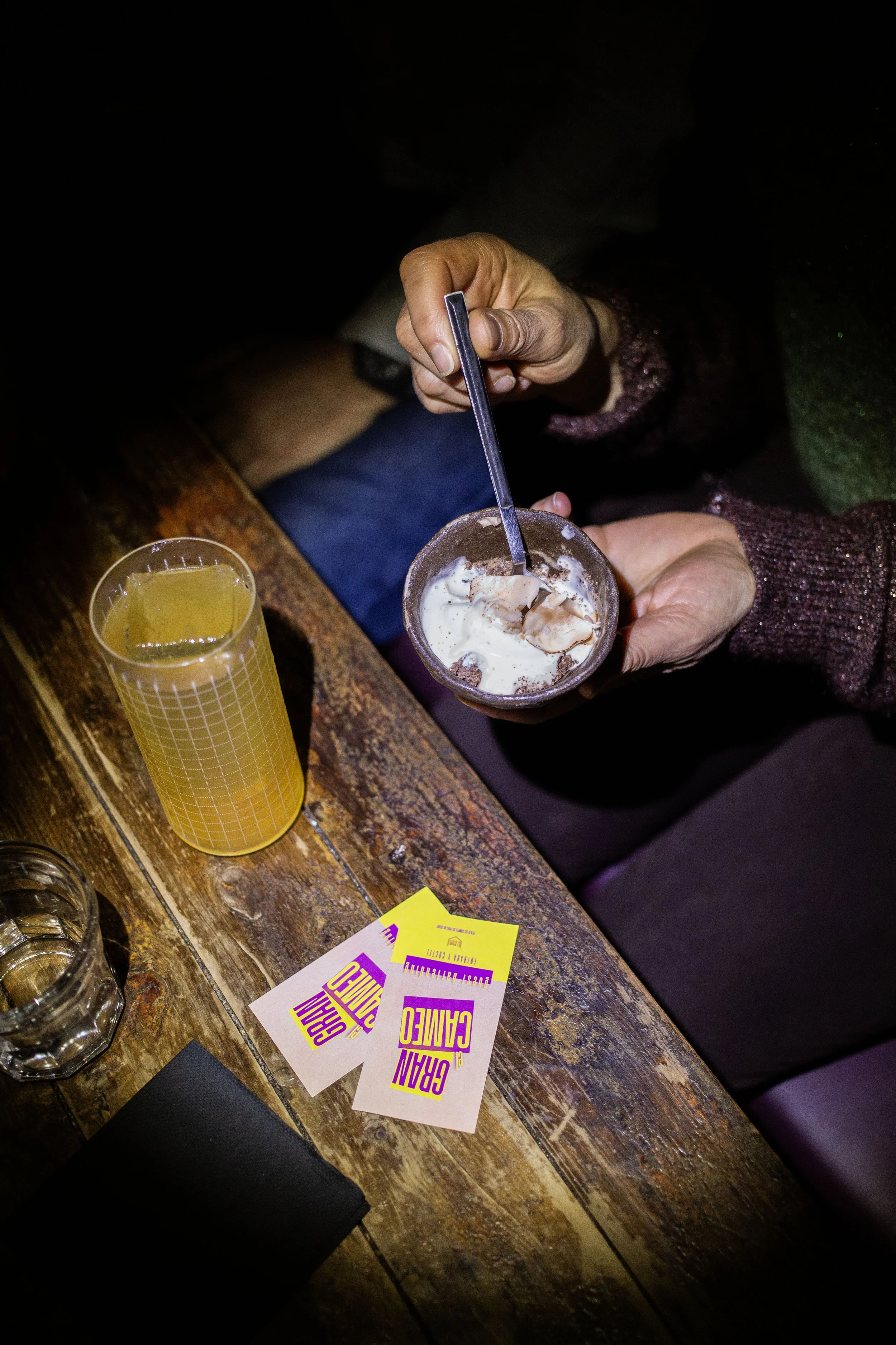 Persona comiendo helado en un restaurante, acompañado de una bebida amarilla y boletos de concierto en una mesa de madera. Contenido de redes sociales.