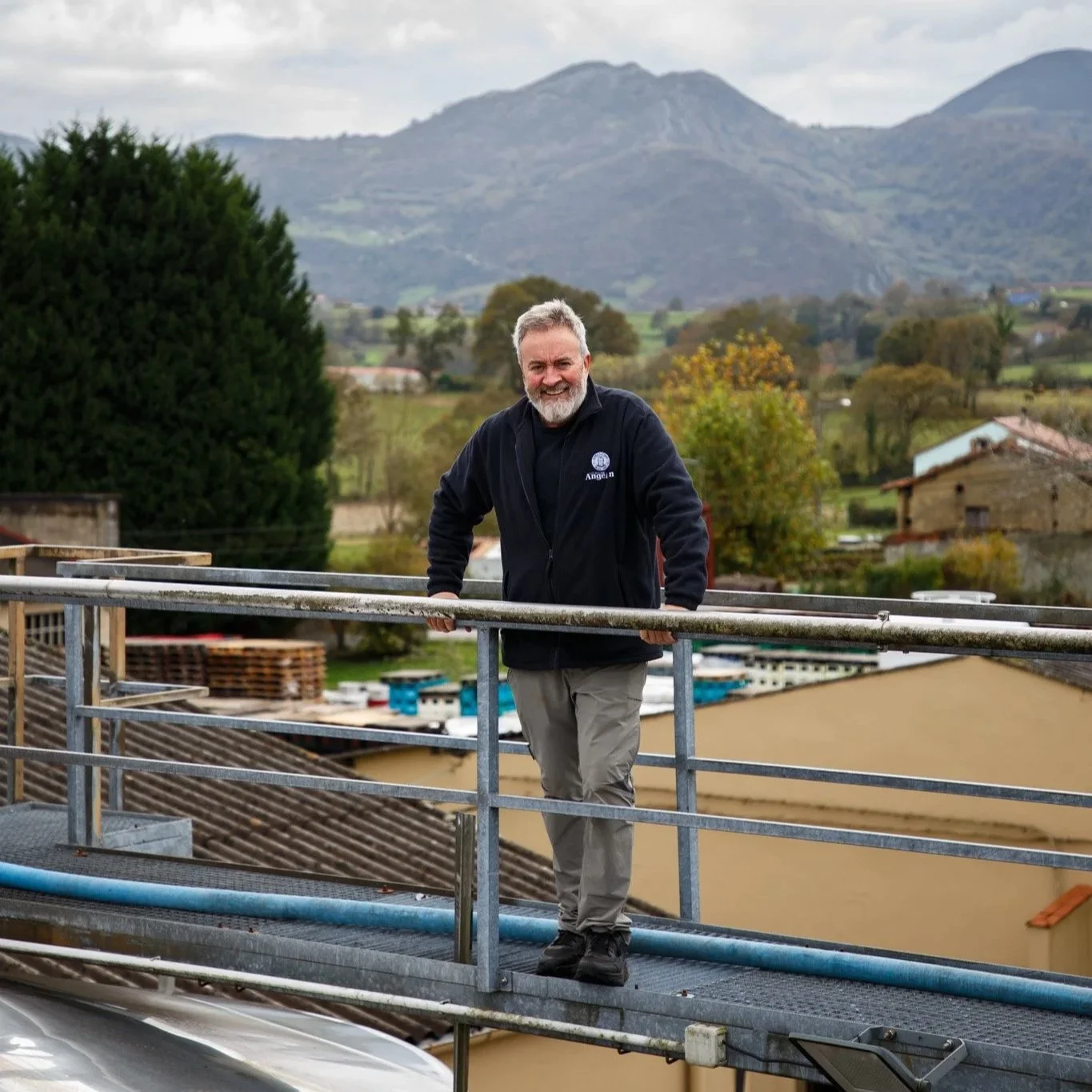 Hombre de cabello y barba gris, sonriendo, de pie en una plataforma metálica con paisaje rural de montañas, árboles y casas en el fondo. Contenido de redes sociales.