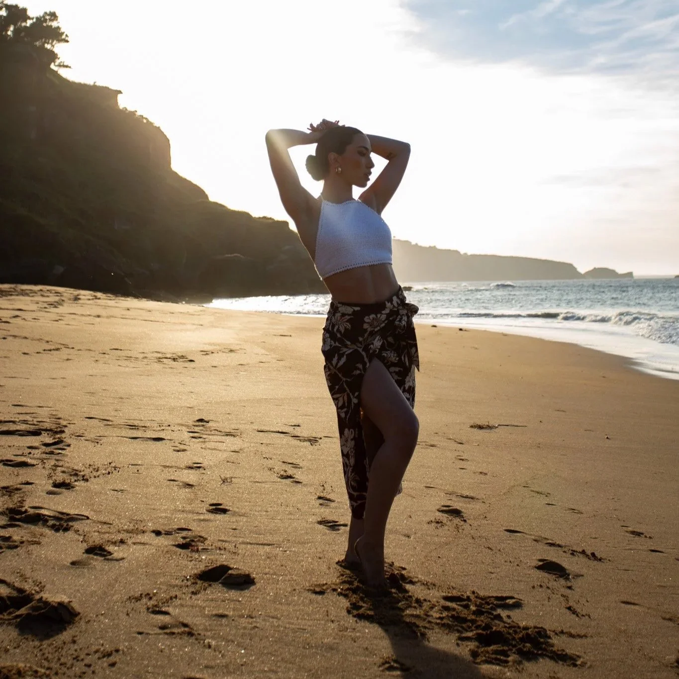 Mujer en la playa durante el atardecer, con un vestido largo y arriba una blusa blanca, posando con las manos en la cabeza frente al mar y colinas en el fondo. Contenido de redes sociales.