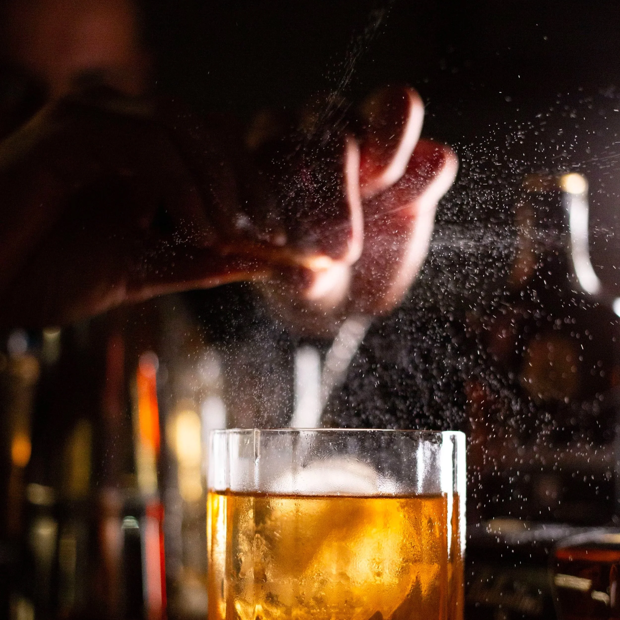 Una mano está colocando hielo en un vaso de bebida con hielo y líquido dorado, en un ambiente con fondo oscuro y desenfocado. Contenido de redes sociales.
