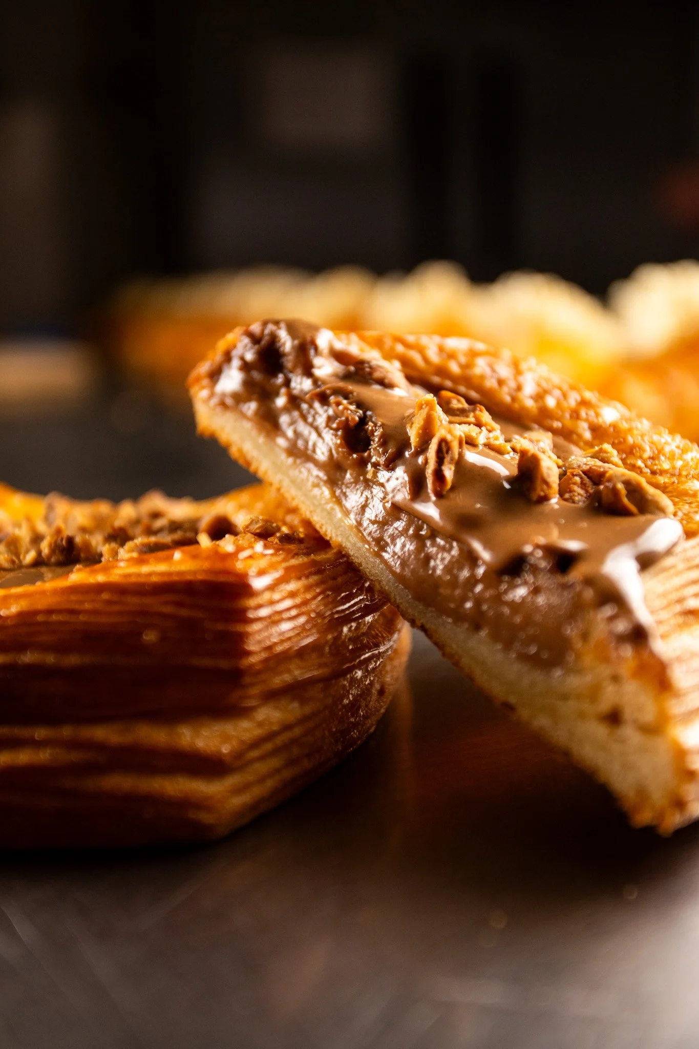 Porción de tarta de dulce de leche con nueces, en primer plano, con más tartas de fondo sobre una superficie oscura. Contenido de redes sociales.